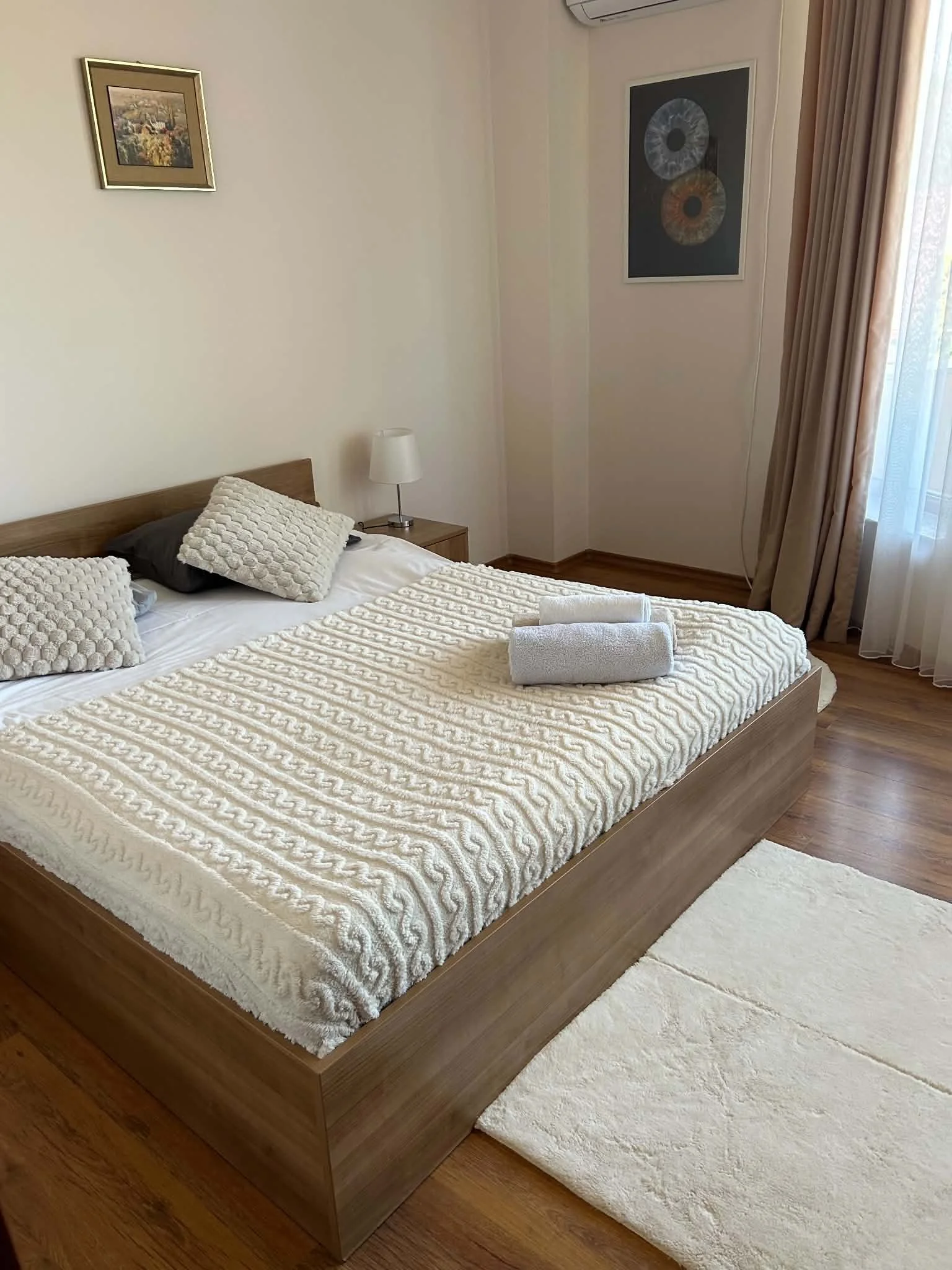 A neatly made bed with cream textured blankets, bath towels, and decorative pillows, in a room with wooden flooring, a nightstand with a white lamp, and framed artwork on the walls, near a window with cream curtains.