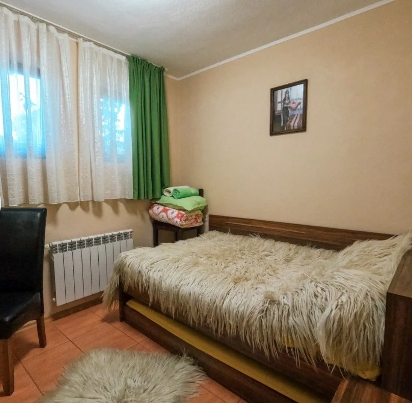 A cozy bedroom with a single bed covered in a fluffy beige blanket, a small table with folded towels, and a black chair. The window has beige and green curtains, and there is a framed picture on the wall.
