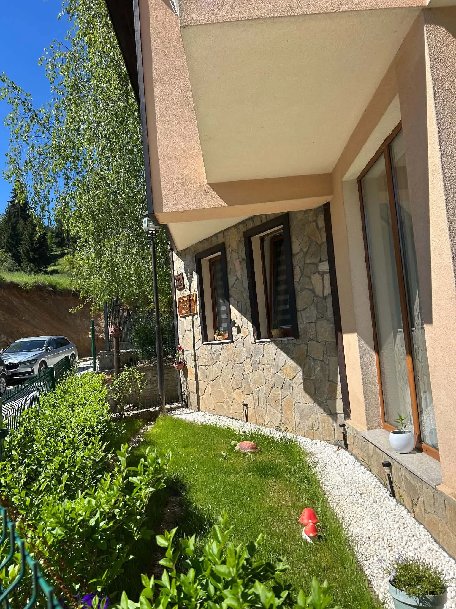 A view of a house with a stone and peach exterior, large windows, a green lush lawn, potted plants, garden decorations, and a car parked in the driveway under a clear blue sky.