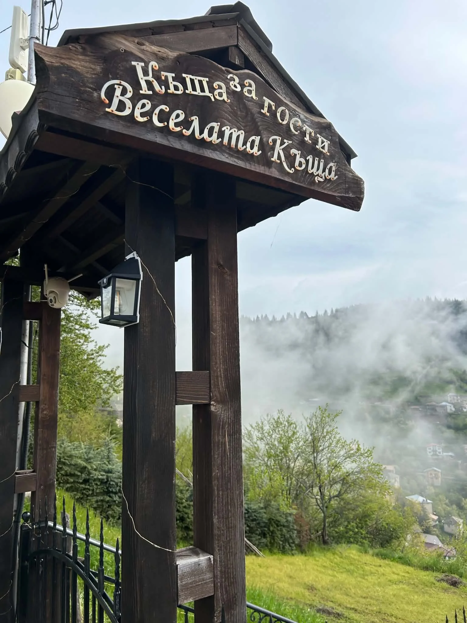 Wooden sign reading 'Къщa за гости ВЕСЕЛАМА Къща' mounted on a wooden structure outdoors with a foggy landscape, trees, and houses in the background.