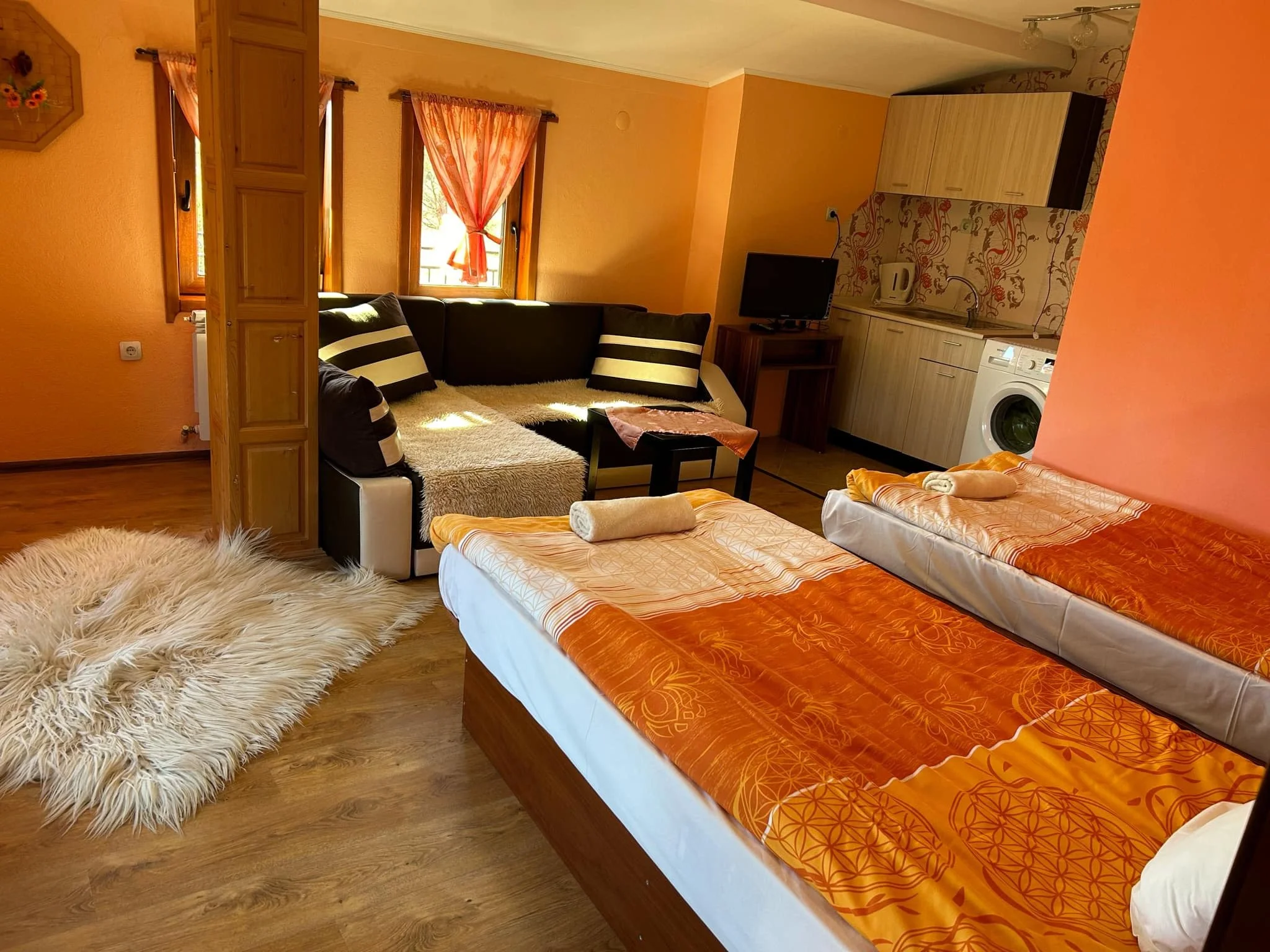 A cozy bedroom with two single beds covered in orange floral bedspreads and freshly folded towels. There is a small flat-screen TV on a wooden stand, a kitchenette with light wood cabinets, a washing machine, a kettle, and a patterned backsplash. Two