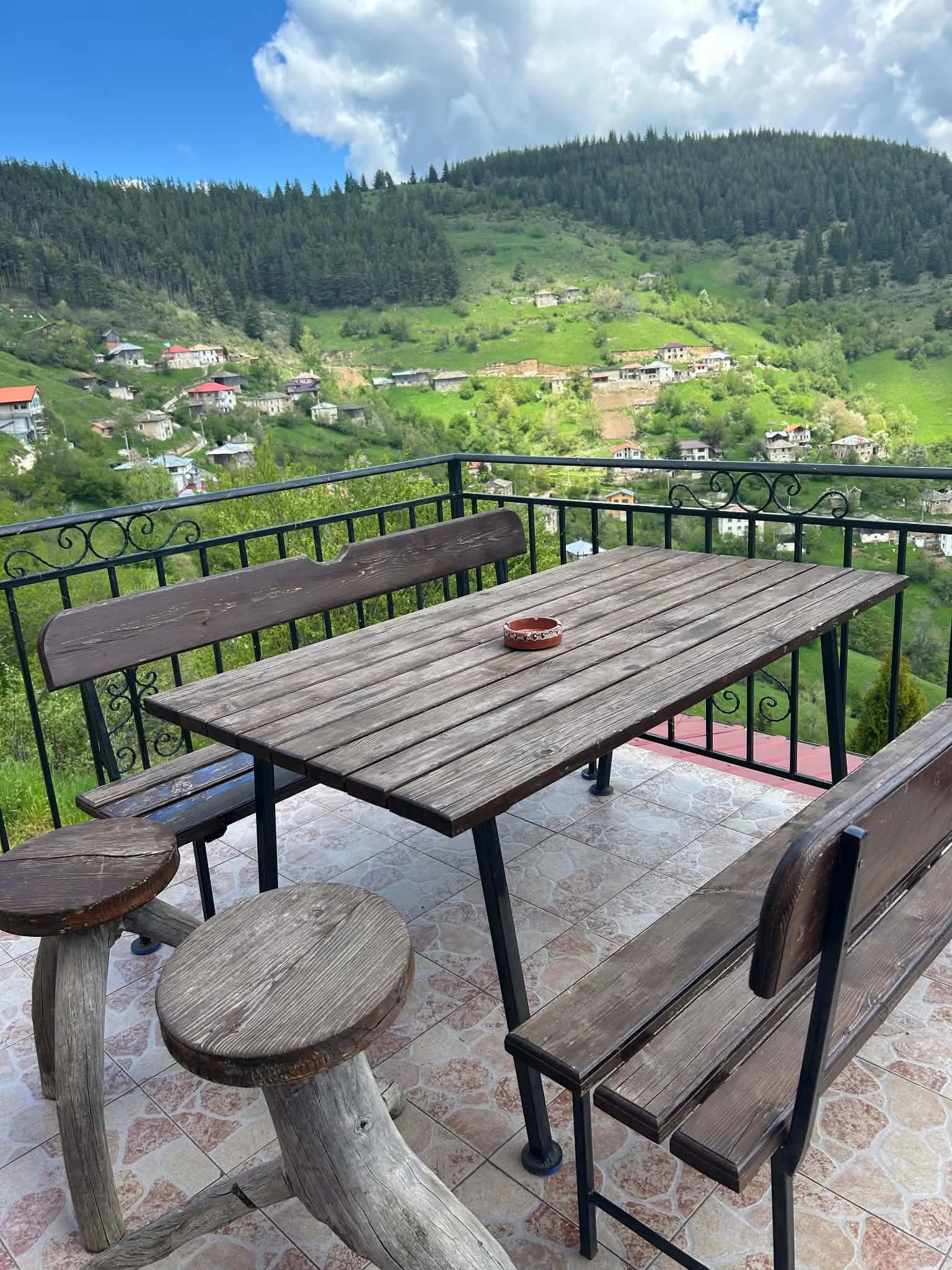 A wooden table and bench set on a tiled balcony overlooking a green hillside with small houses and dense forest under a partly cloudy sky.