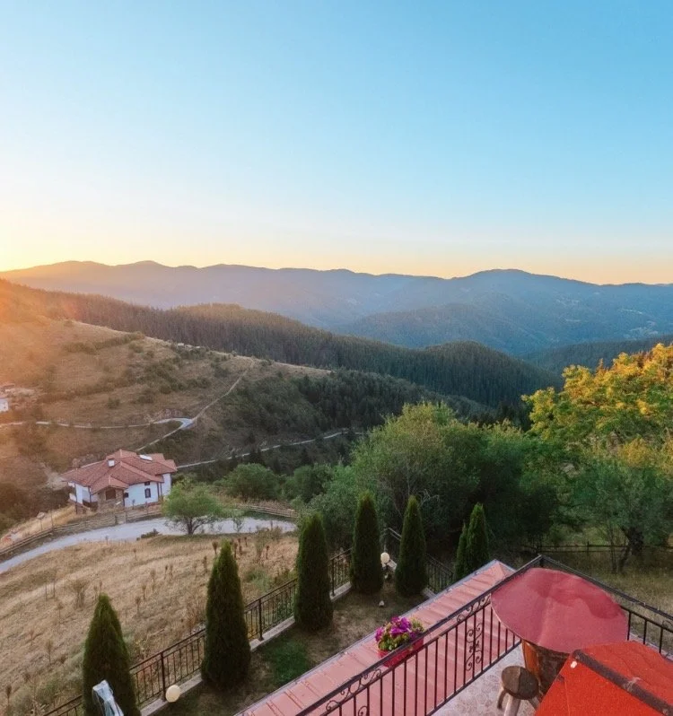 Scenic view of mountains at sunset with a balcony in the foreground, featuring a red umbrella, small table, and potted flowers.
