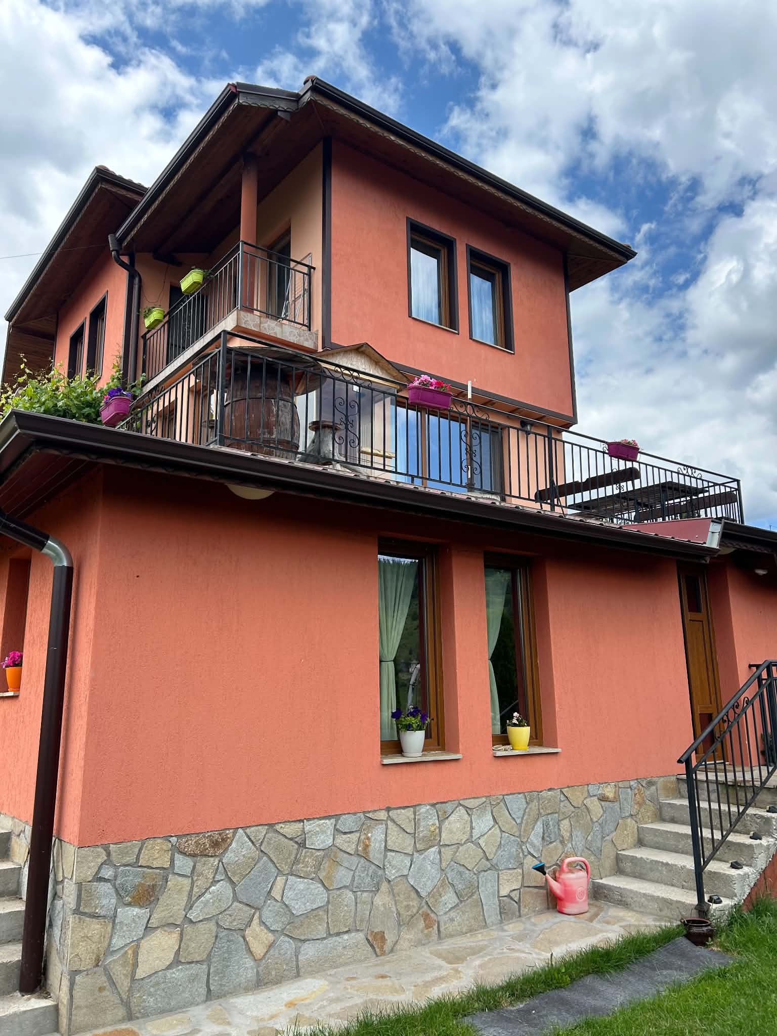 A two-story house with a pink exterior, stone foundation, and a small balcony with flower pots. There are windows with green curtains and outdoor plants. The sky is partly cloudy.