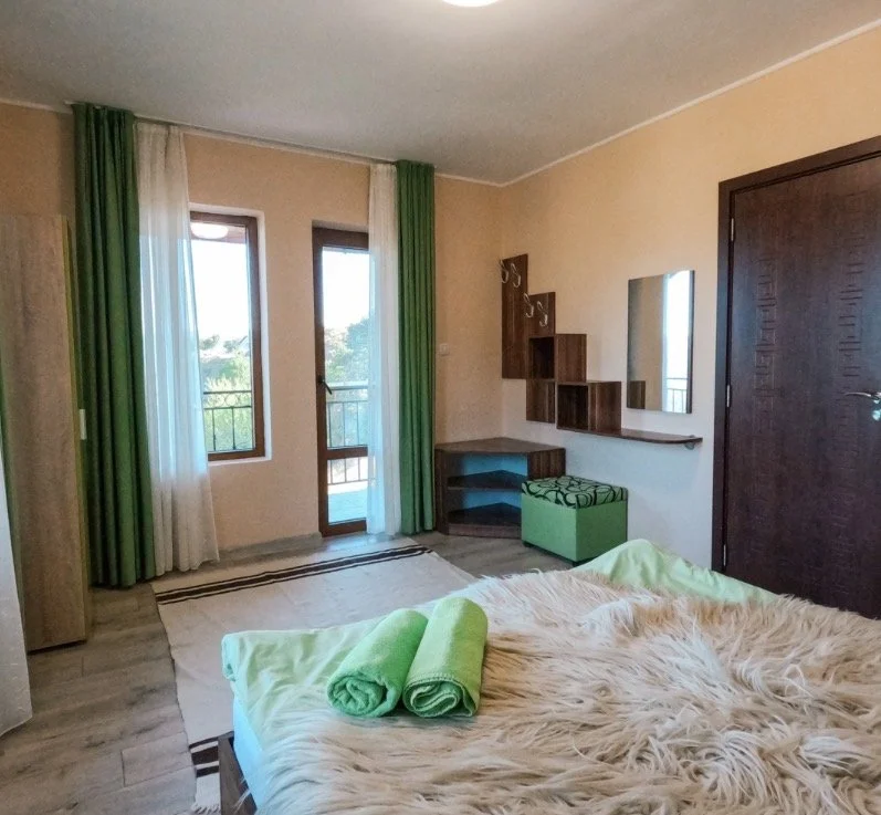 A bedroom with beige walls, a bed with a furry blanket, two green towels, and green curtains. There are two glass doors leading to a balcony, a wooden wardrobe, and a mirror on the wall.