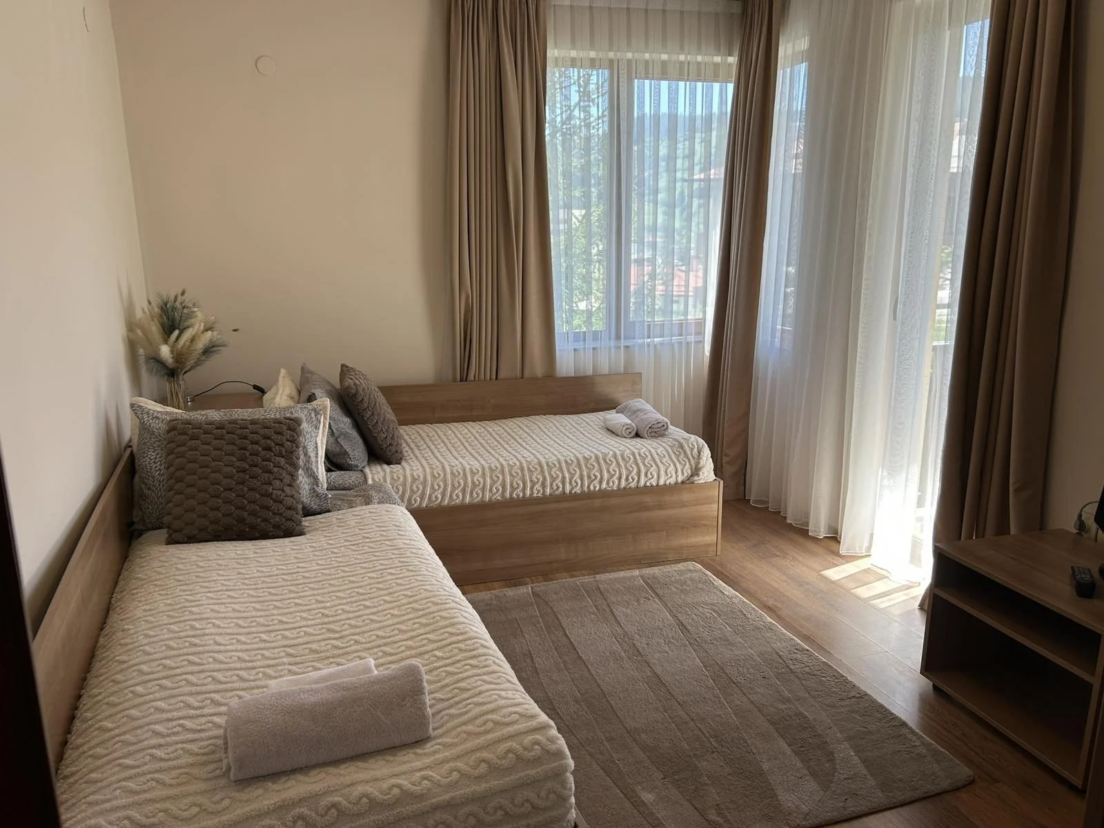 A bedroom with two beds, one on the left with a textured blanket and pillows, and a bed against the window with rolled towels on top, light-colored curtains,:one window, and wooden furniture, including a small TV stand.