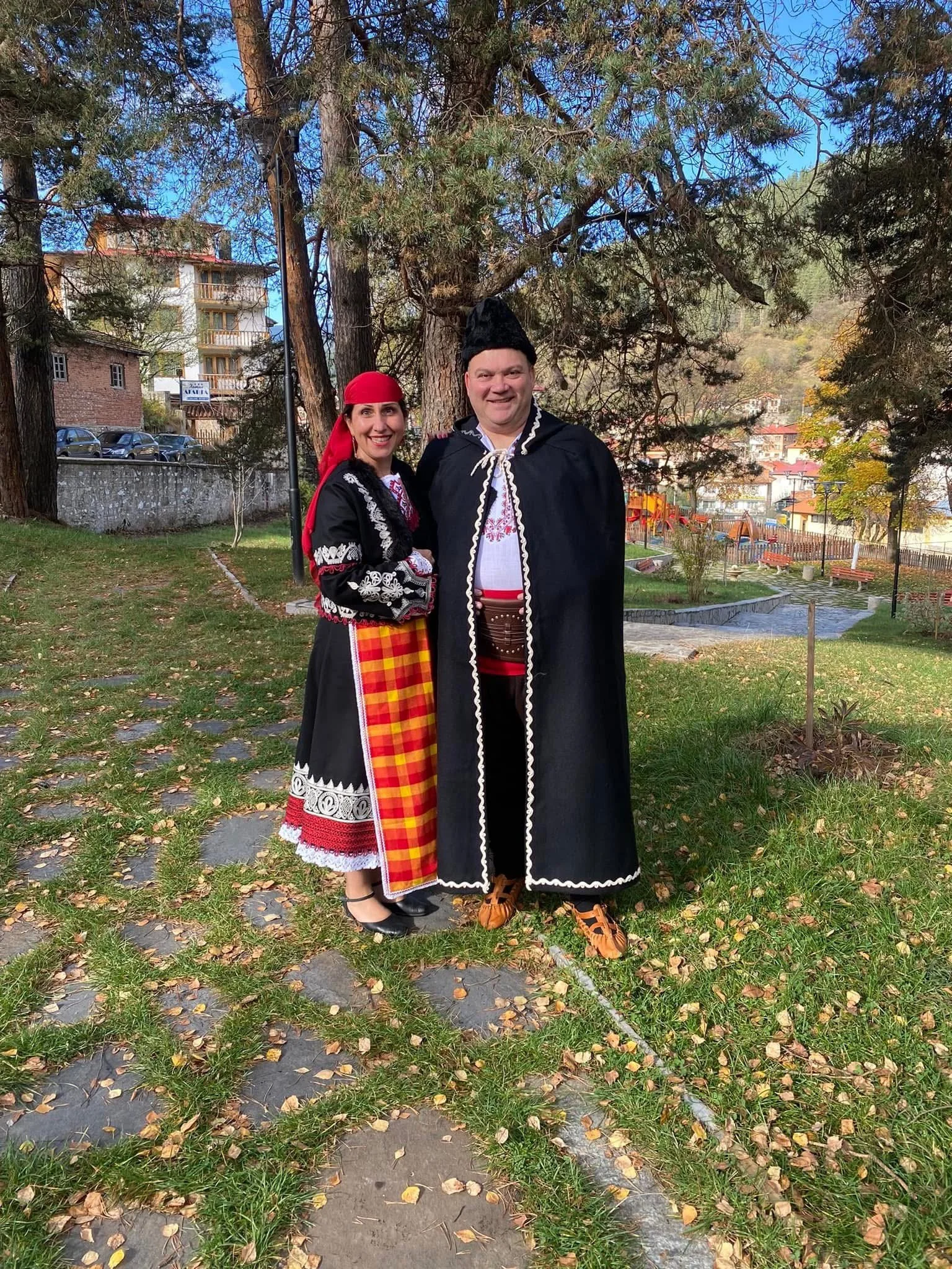 Two people dressed in traditional Eastern European folk costumes standing outdoors in a park with trees and buildings in the background.