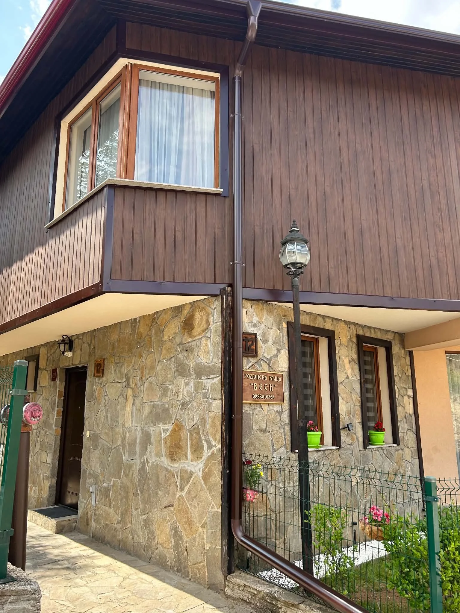 A stone house with a wooden balcony, a lamp post, and potted plants on the windowsills.