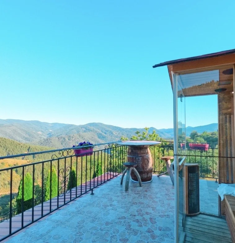 Balcony with a view of green mountains and a clear blue sky, decorated with a small table and two stools, enclosed by a glass railing, attached to a wooden house.