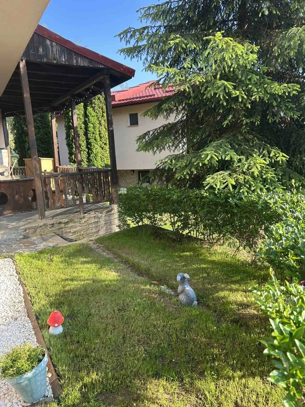 A backyard garden with green grass, small shrubs, and a large evergreen tree. There is a statue of a rabbit and decorative mushrooms on the lawn. A wooden porch with a roof and hanging lights is visible on the left, with a stone pathway leading to it