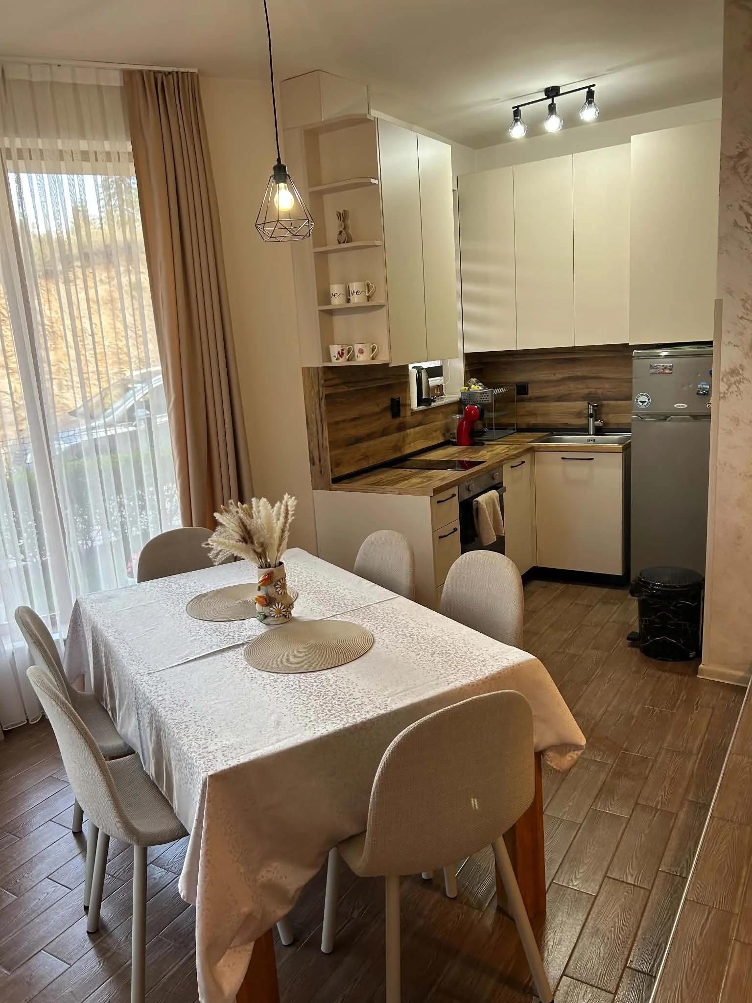 A kitchen and dining area with a dining table covered with a white tablecloth, surrounded by beige chairs, a vase with dried flowers on the table, built-in cabinets, a countertop with appliances including a coffee maker and a red espresso machine, a 