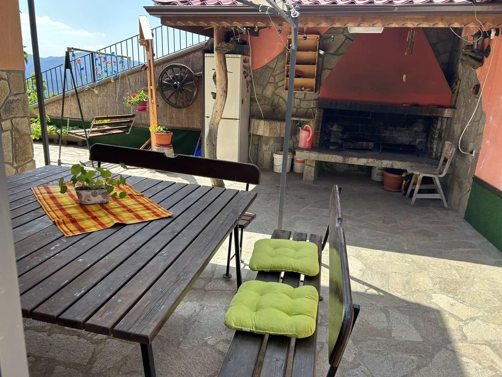 A covered outdoor patio with a wooden dining table with a checkered tablecloth, potted plants, and metal chairs with bright green cushions. In the background, there is a fireplace, a refrigerator, and a swing with colorful lights and mountain views.