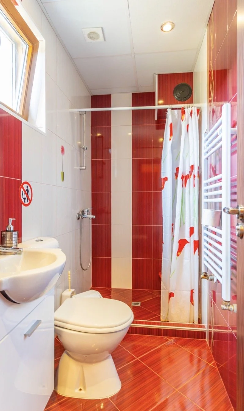Small bathroom with red and white tiled walls and floor, a toilet, a sink with soap dispenser, a window, and a shower area with a curtain decorated with red flowers.
