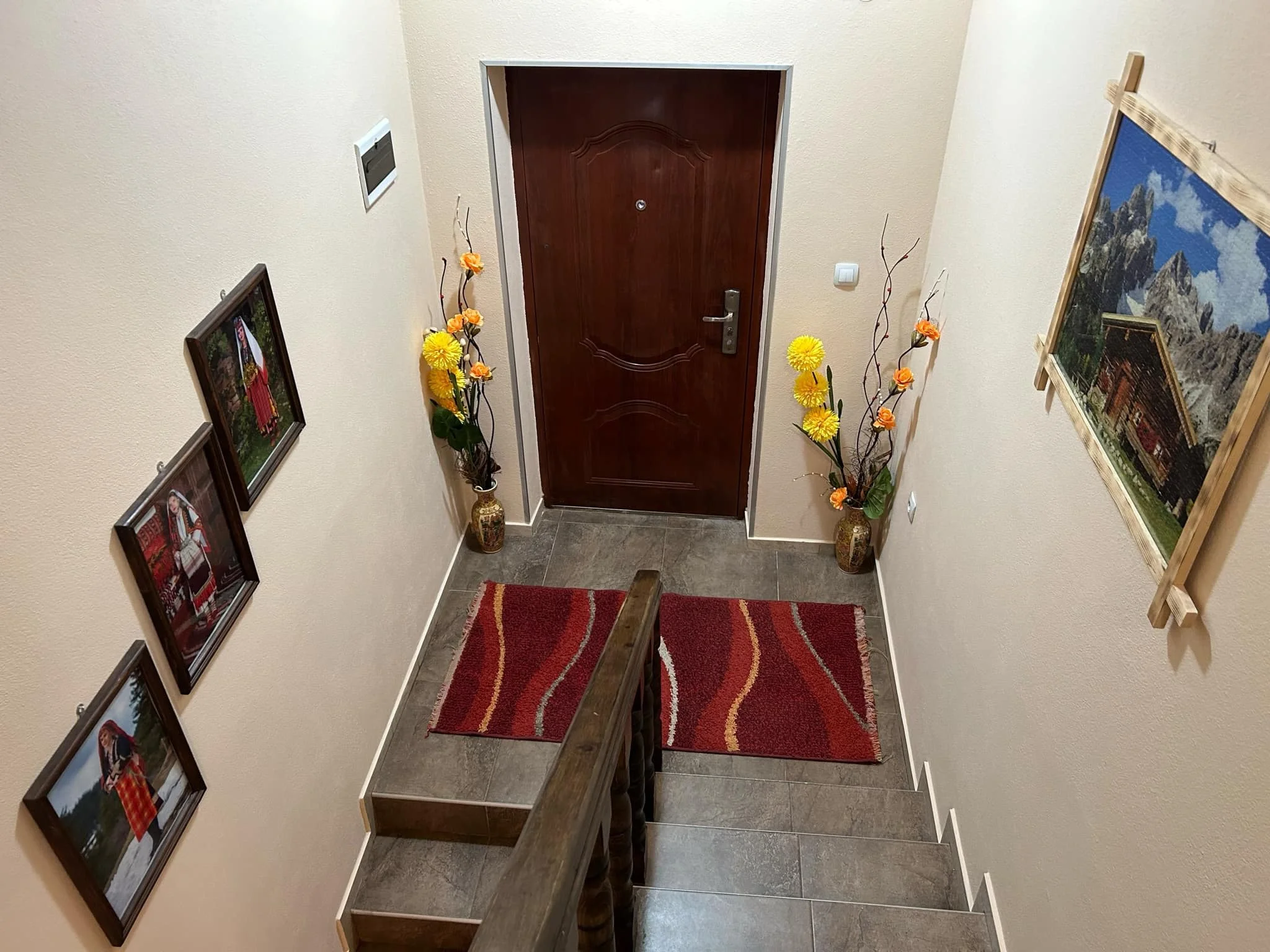 A view of an entryway with a wooden front door, flanked by two tall vases with yellow and orange artificial flowers. On the left wall, there are three framed photographs. To the right, there is a landscape painting on the wall. The floor is tiled wit