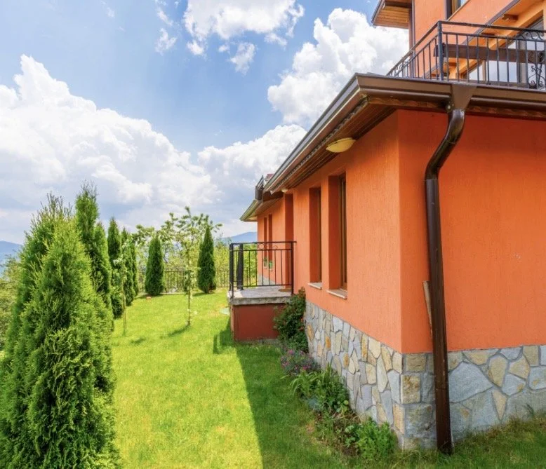 Side view of an orange house with a stone foundation, a small balcony, and a garden with tall green trees and grass, under a partly cloudy sky.