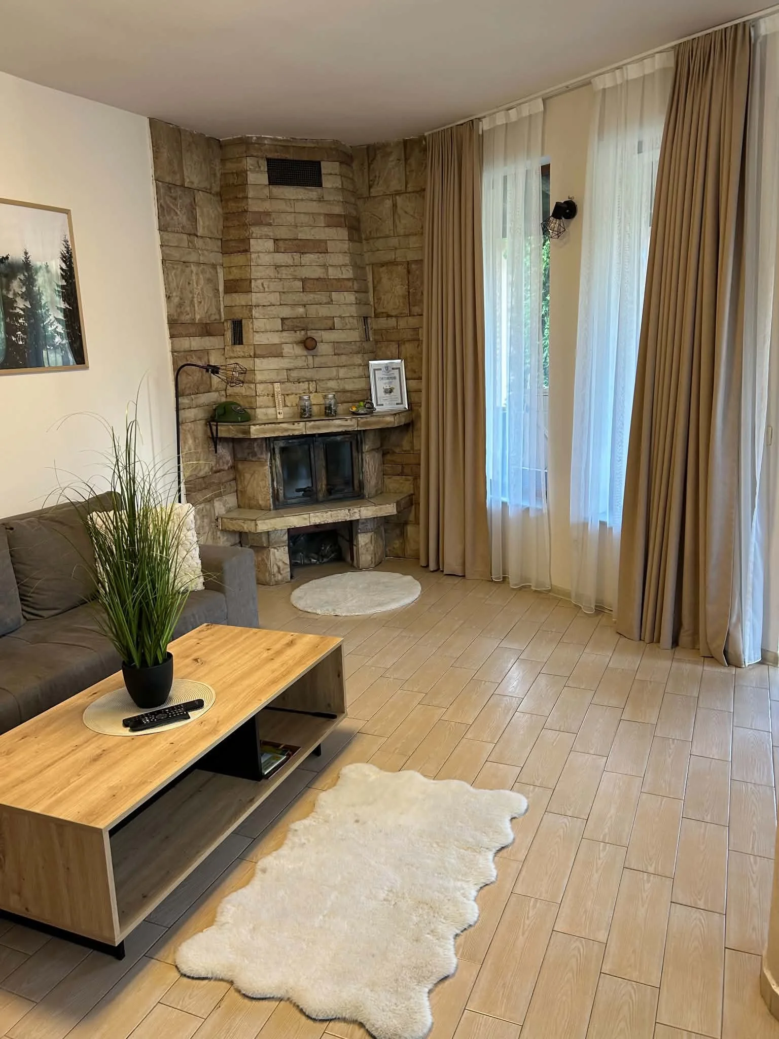 Living room with beige tiled floor, a gray sofa, a wooden coffee table with remote controls, a cream sheepskin rug, a brick fireplace, gold curtains, and a window with sheer white drapes.
