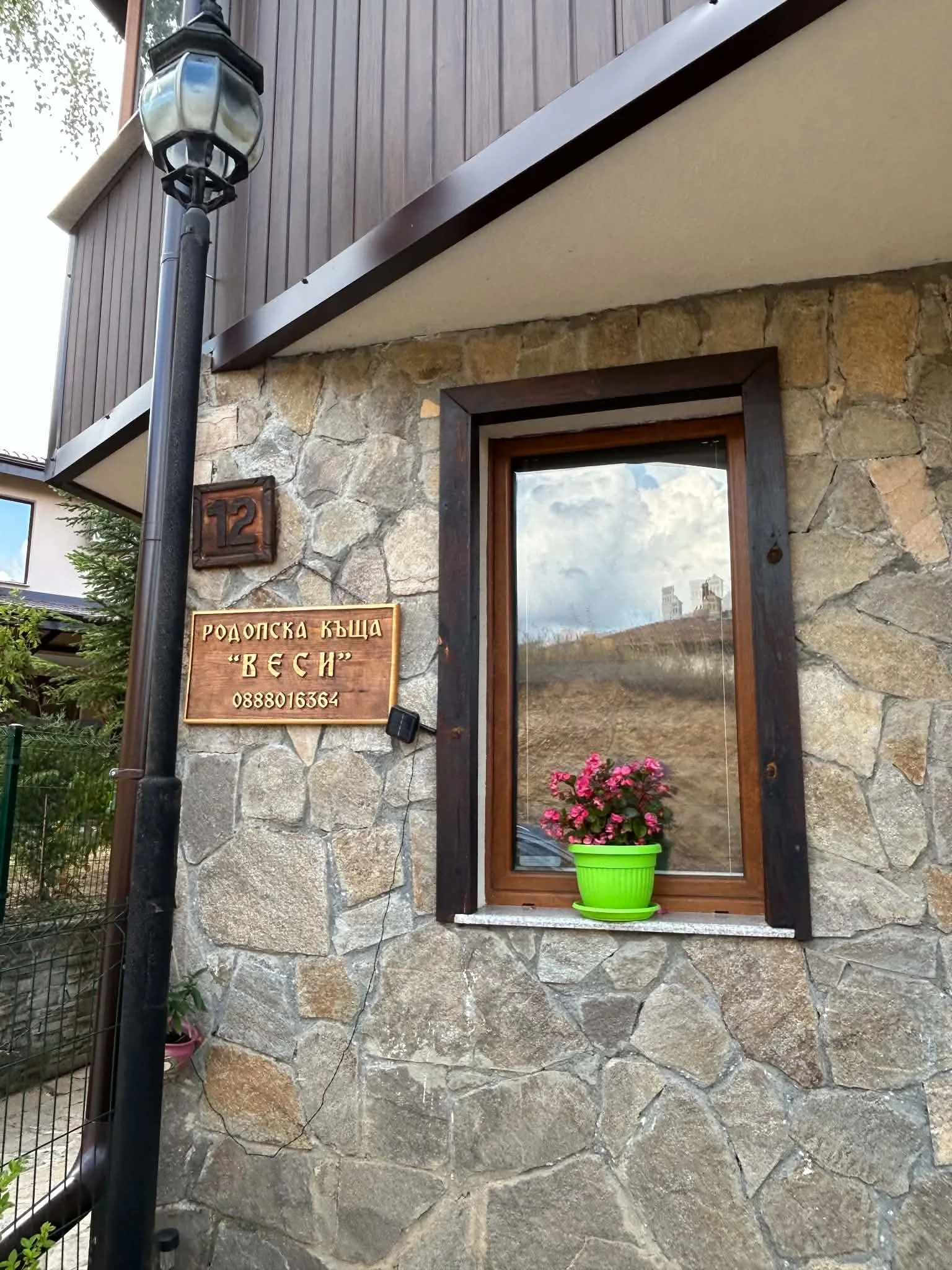 A stone wall with a wooden window frame and a potted pink flower plant on the windowsill. There is a wooden sign with text and a house number plaque mounted on the wall. A black lamp post is visible in the foreground.