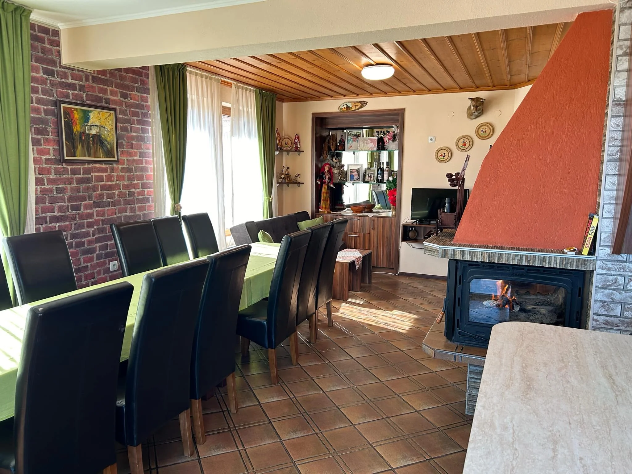 A cozy dining room with black leather chairs around a table covered with a green tablecloth. There is a brick wall on one side and a window with green and white curtains. In the corner, there is a fireplace with a fire burning, and decorative items a