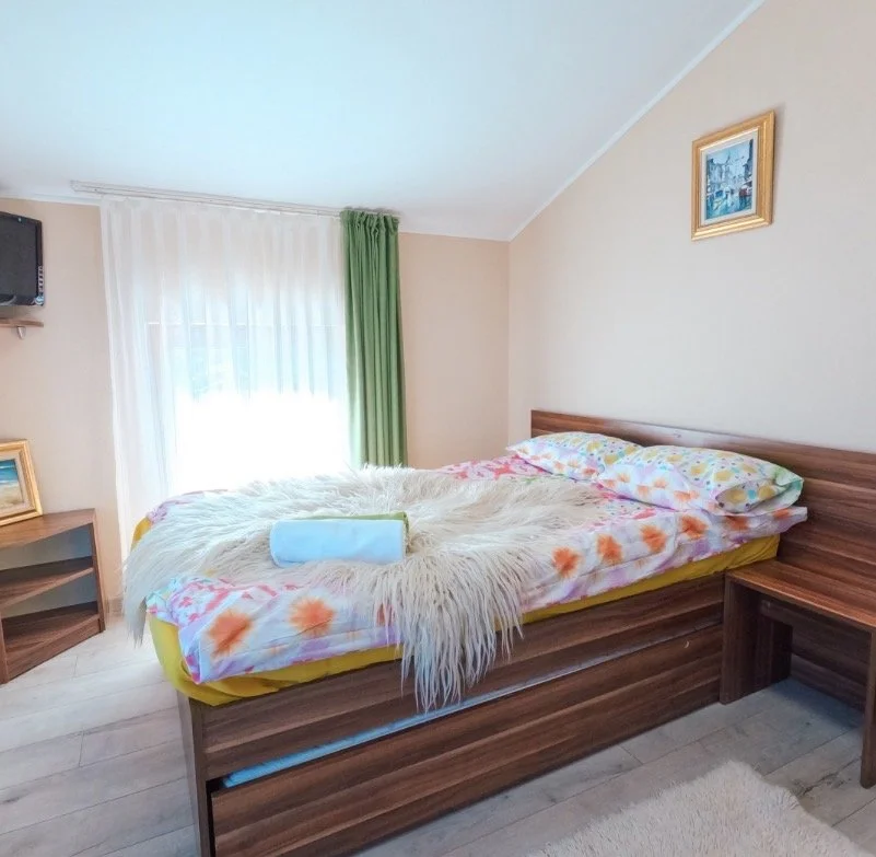 A cozy bedroom with a wooden bed frame, floral sheets, a fluffy white throw blanket, and a rolled towel. There's a window with white and green curtains, a small wooden nightstand with a framed picture, and a corner-mounted TV. A picture hangs on the 