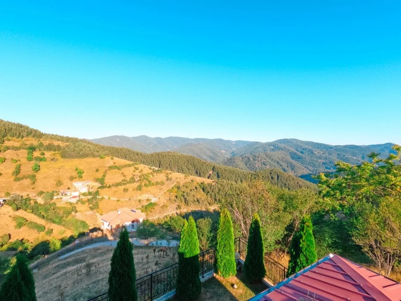 Scenic view of green mountains and hills under a clear blue sky, with a foreground featuring a red-tiled roof, tall green trees, and a private fenced yard.