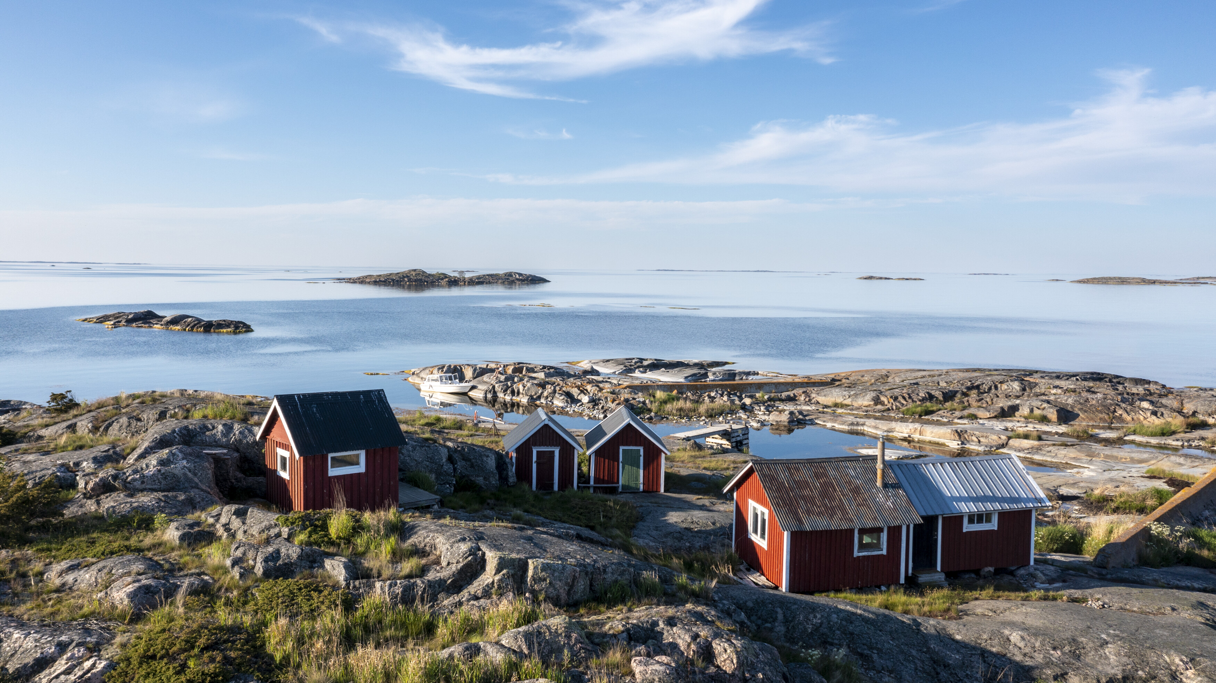 Coastal scene with red houses, rocky shoreline, calm blue water, and small islands under a partly cloudy sky.