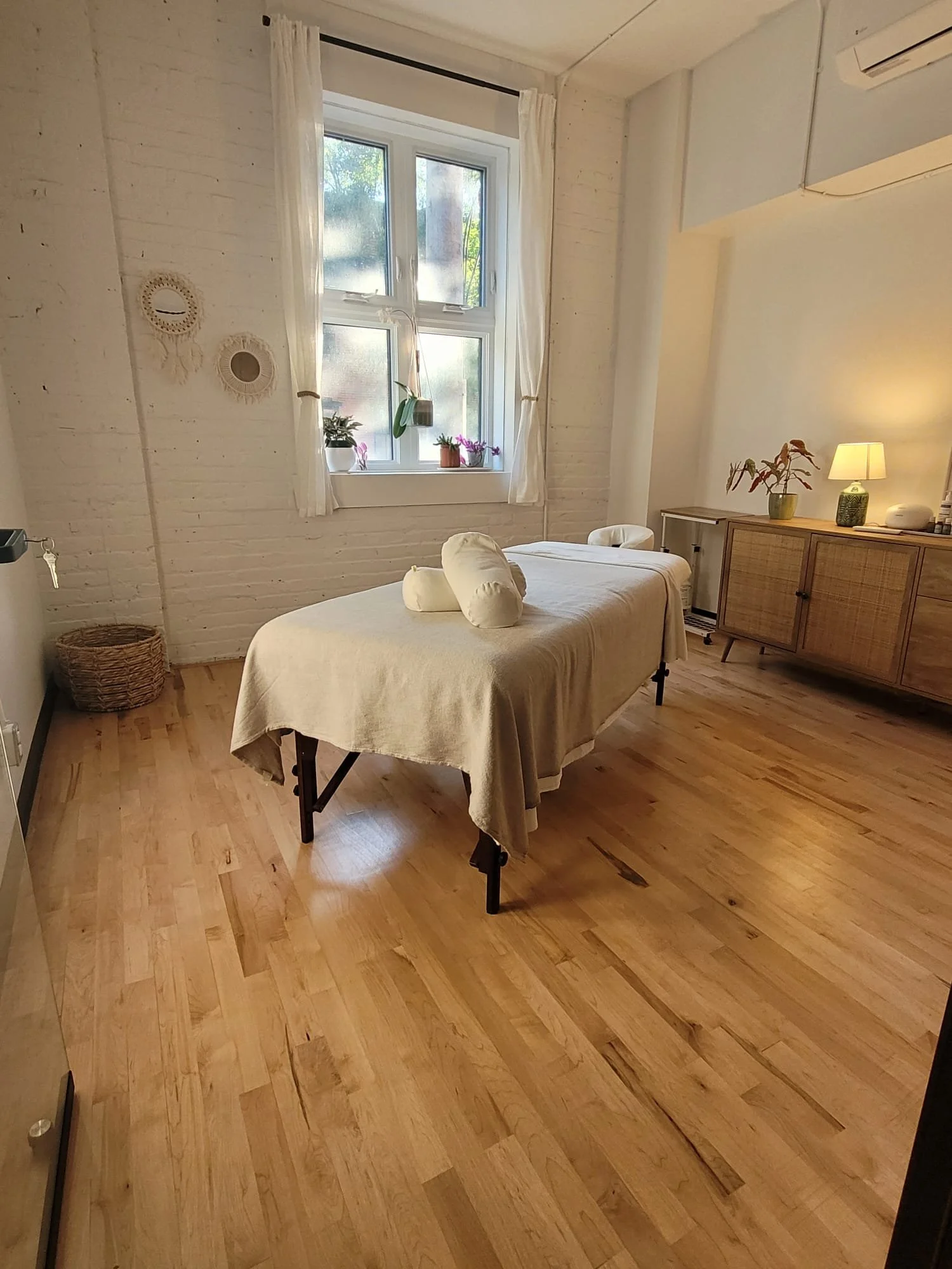 A massage table with a cream-colored sheet and pillow, set in a cozy room with wooden floors, a large window with white curtains, a sideboard with a lamp, plants, and decorative objects.