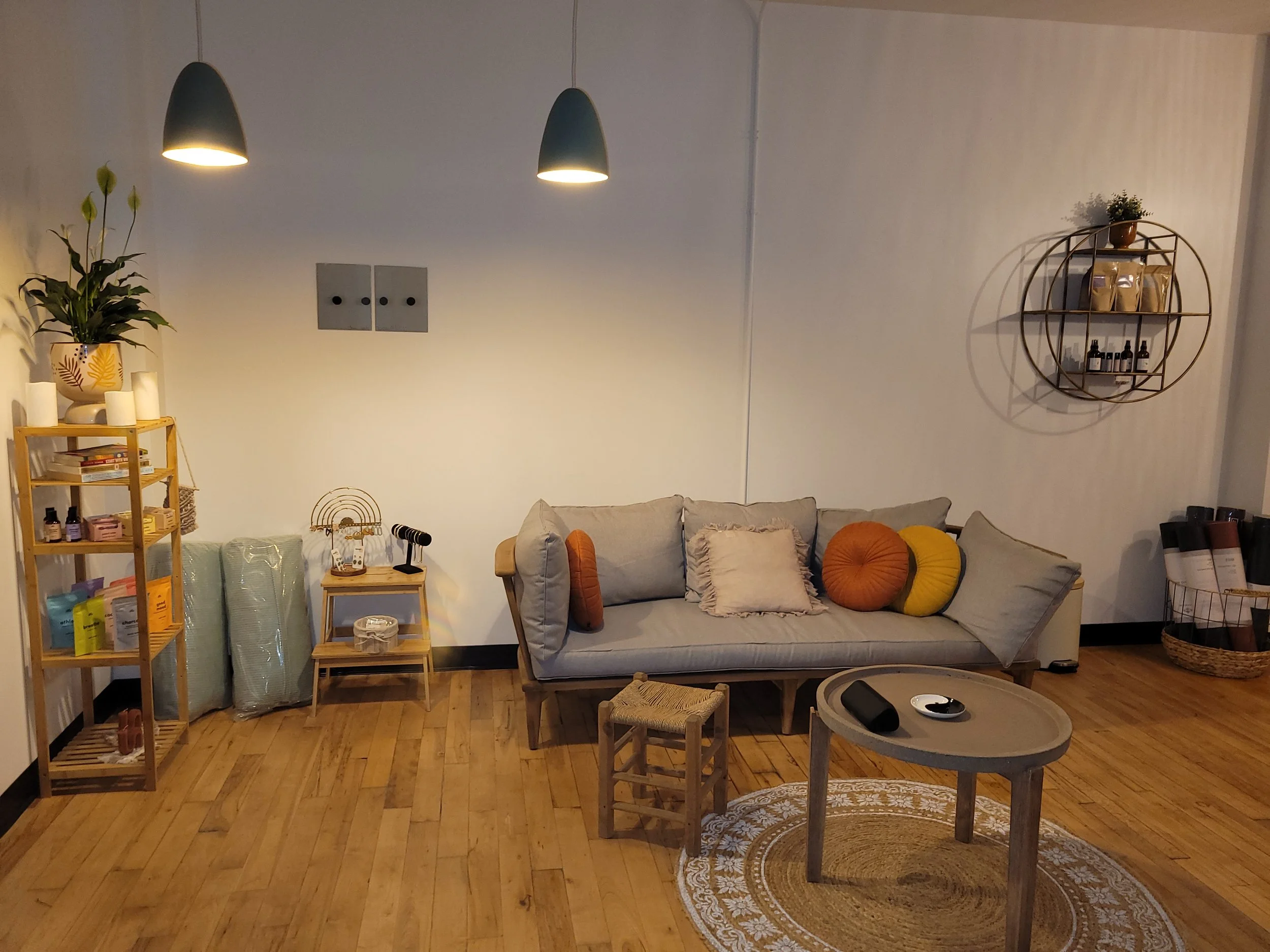 Living room with wooden flooring, featuring a light gray sofa with velvet and textured pillows, circular rug, coffee table with electronic devices, minimalist wooden shelves with various items, hanging pendant lights, and decorative plants.