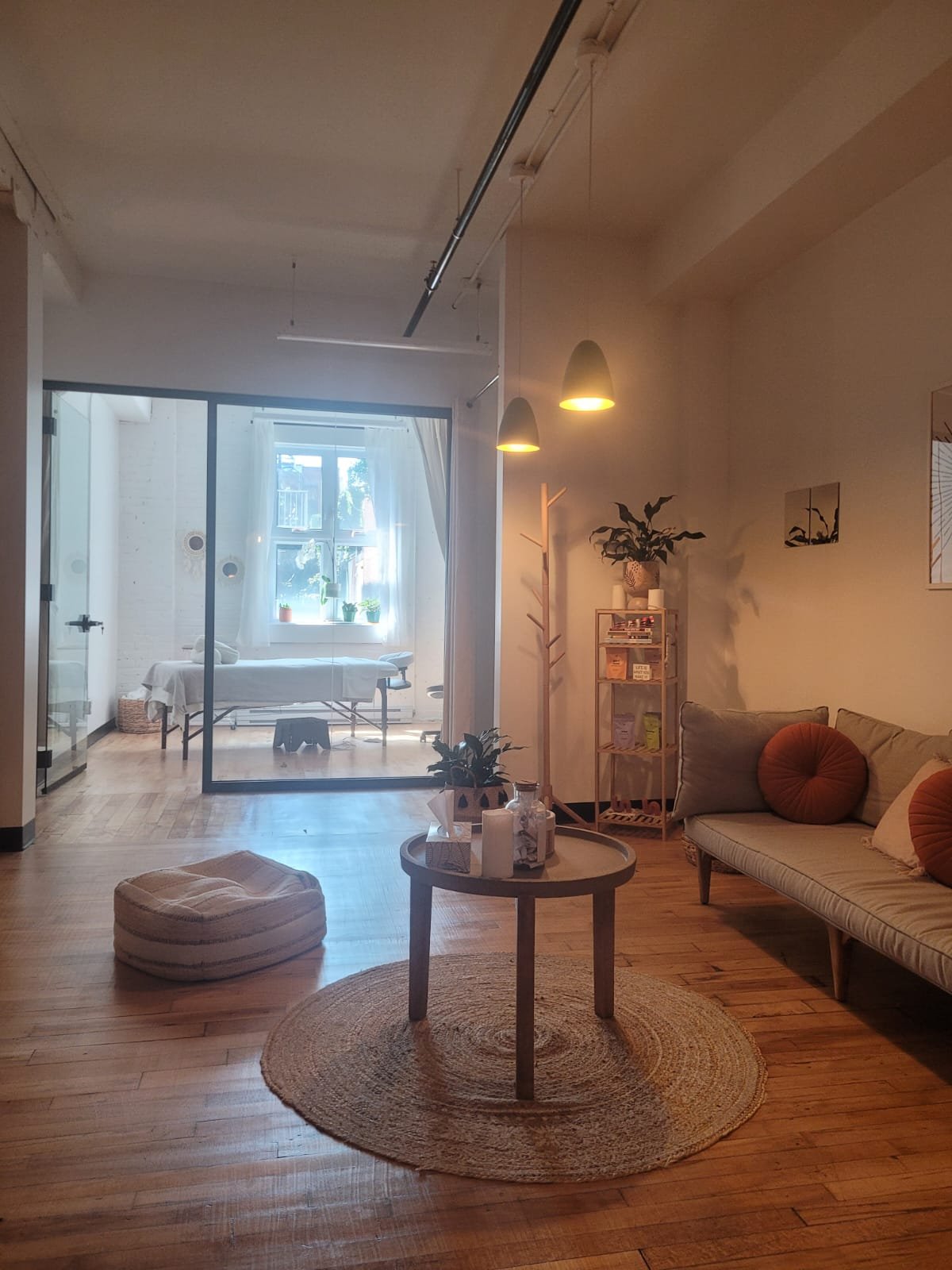 Living room with wooden floor, beige sofa with round pillows, small round coffee table with plants, candles, and tissue box, standing coat rack, bookshelf, and framed art on the wall, with a separate room visible through a glass door.