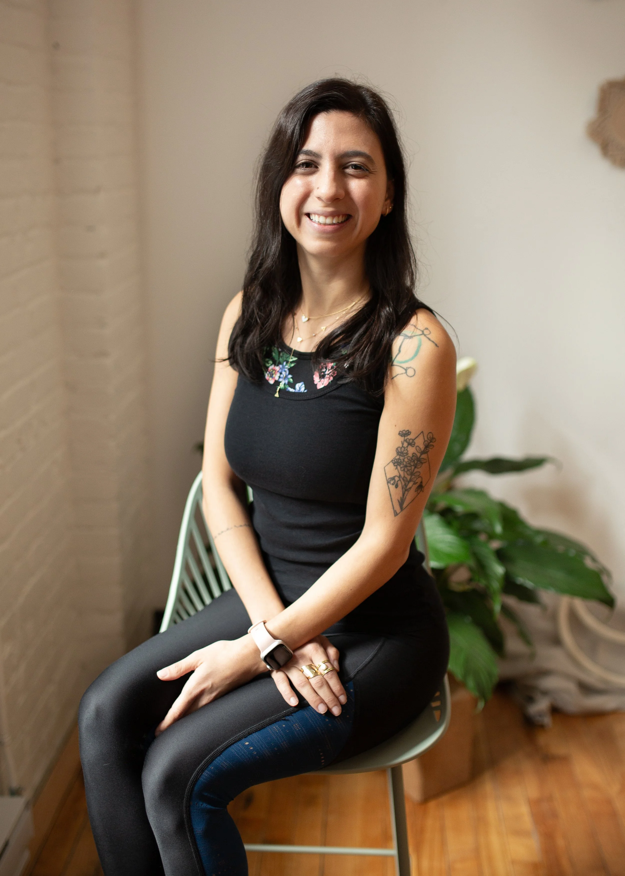 A woman with dark brown hair, wearing a black sleeveless top with floral embroidery and black pants, sitting on a gray chair in front of a beige wall and a potted green plant, smiling at the camera.