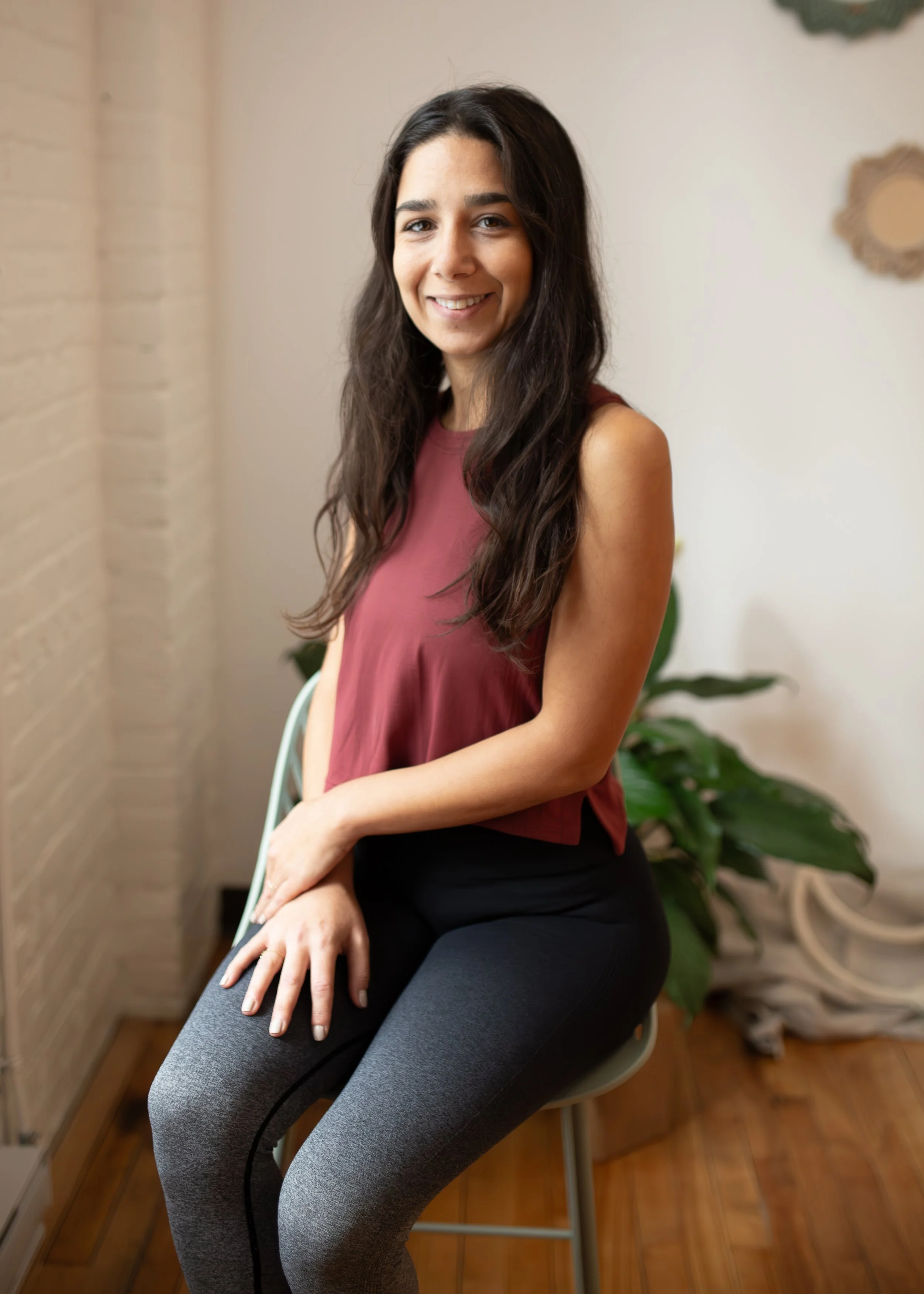 A young woman with long dark hair smiling, sitting indoors on a white chair in front of a white wall with a decorative mirror and green plant, wearing a sleeveless maroon top and black leggings.