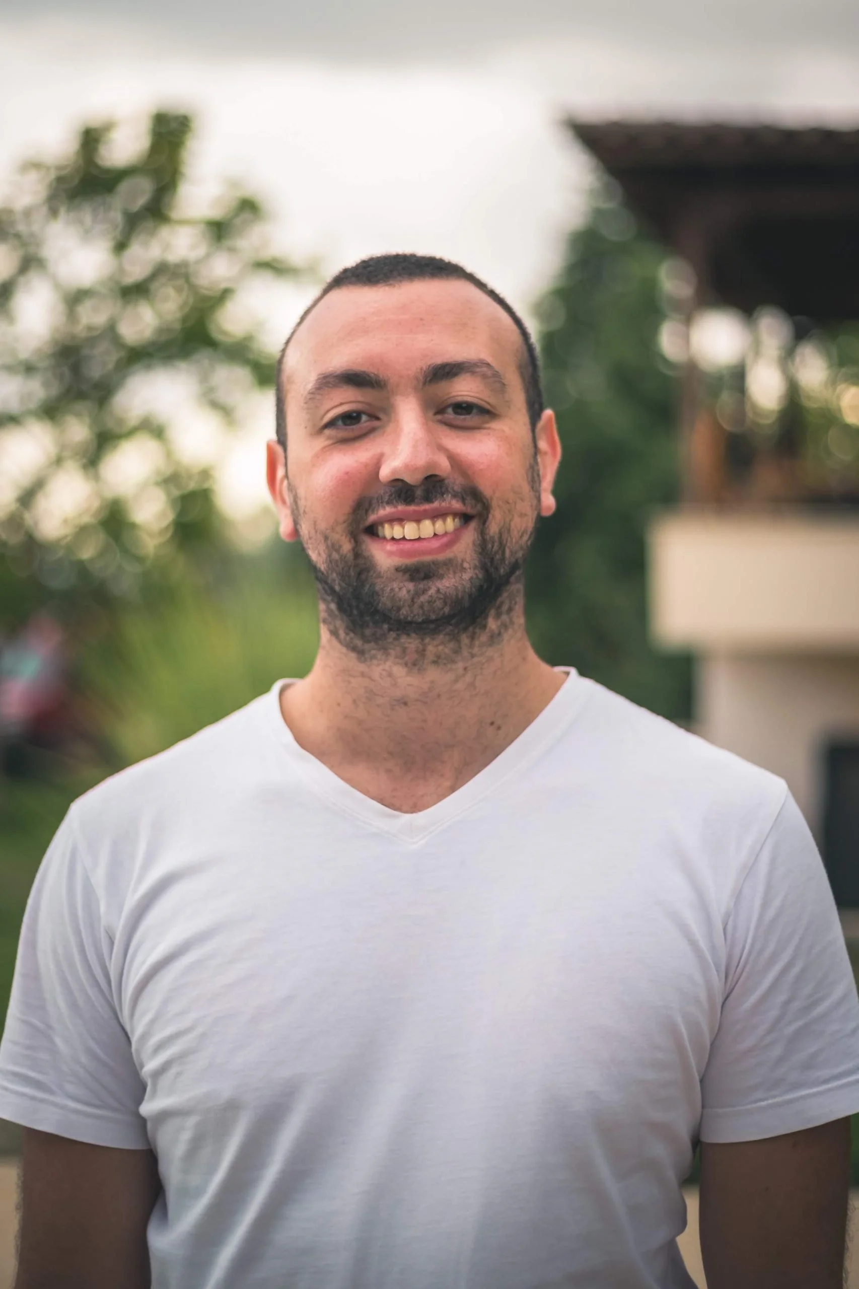 A smiling man with short dark hair, beard, and mustache, wearing a white V-neck T-shirt, standing outdoors with blurred green trees and a structure in the background.