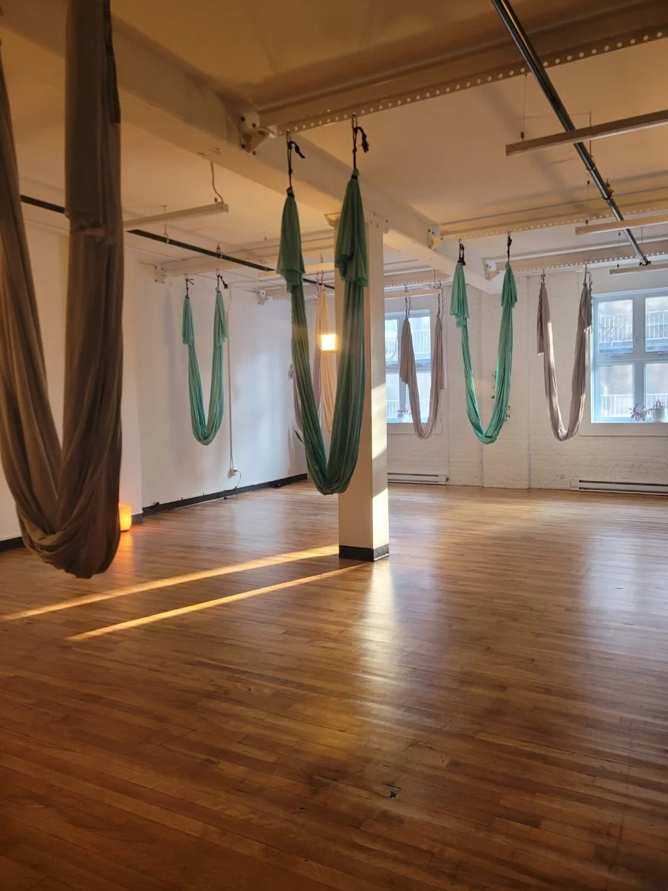 Empty yoga or aerial fitness studio with wooden floors and hanging aerial hammocks in various shades of green and beige, illuminated by natural light from large windows.