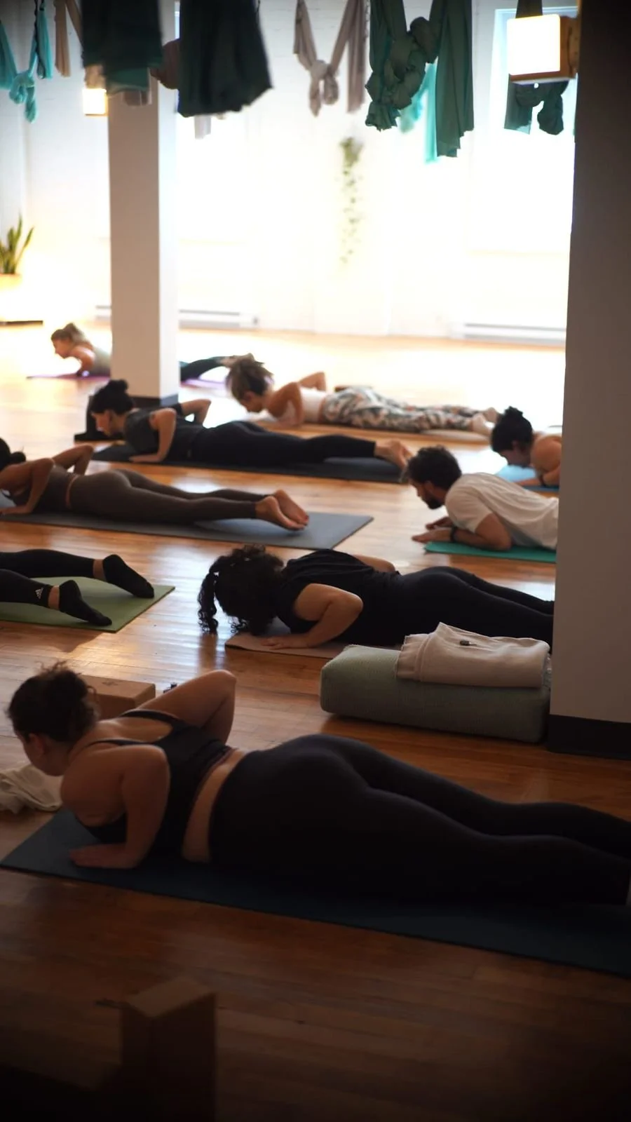 People practicing yoga in a bright studio, lying on yoga mats in a plank position with their arms bent, wearing athletic clothing.
