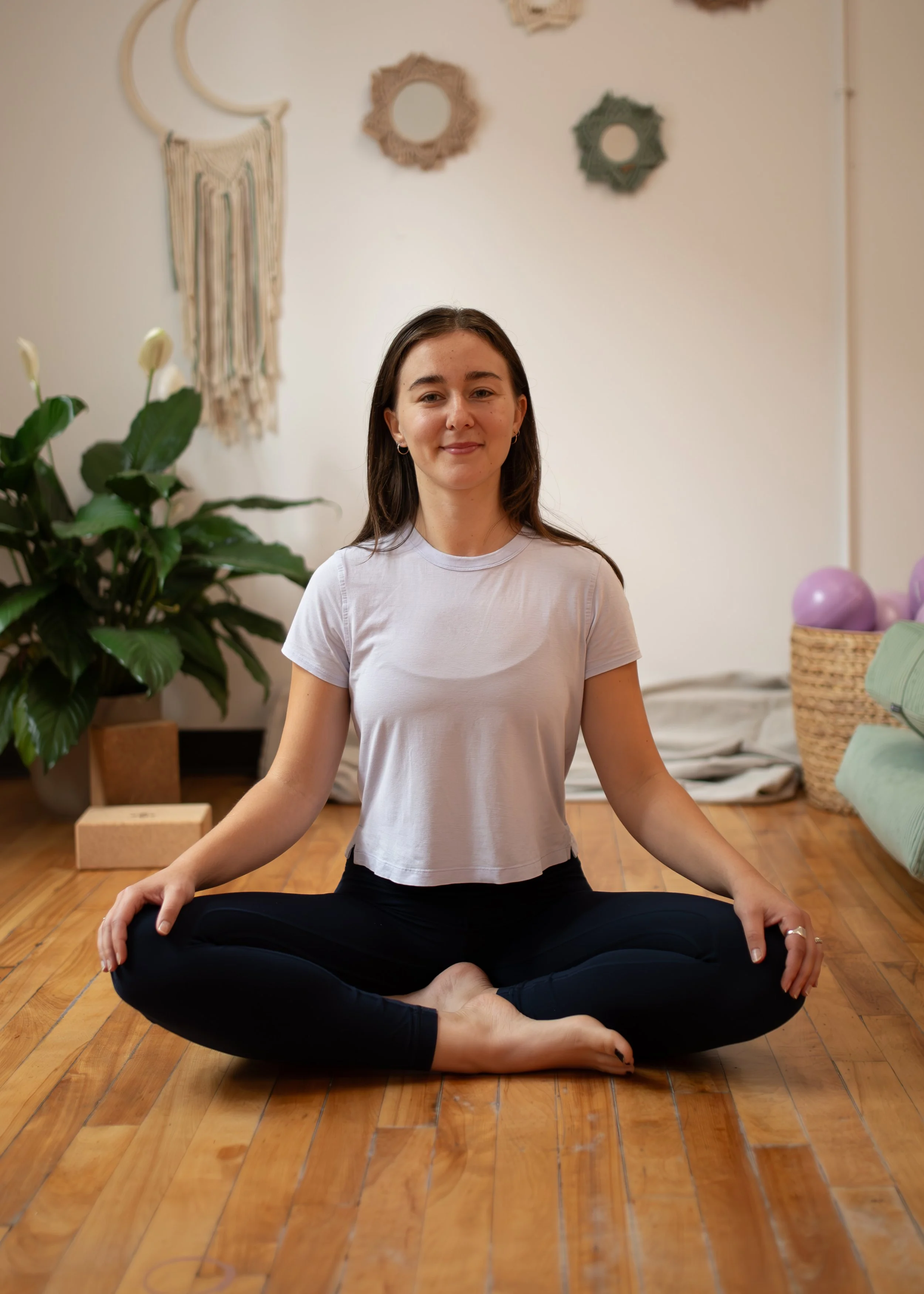 A woman practicing yoga or meditation in a relaxed cross-legged seated pose indoors, with a calm expression. She is wearing a white T-shirt and black pants. The background includes decorative wall hangings and a basket of purple balloons, with wooden flooring and some green plants.