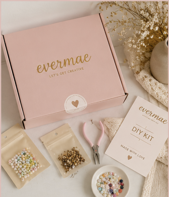 Pink box labeled 'evermae Let's Get Creative' next to DIY kit supplies including colorful beads, small golden charms, pink pliers, and instruction sheet, with a vase of dried flowers in the background.