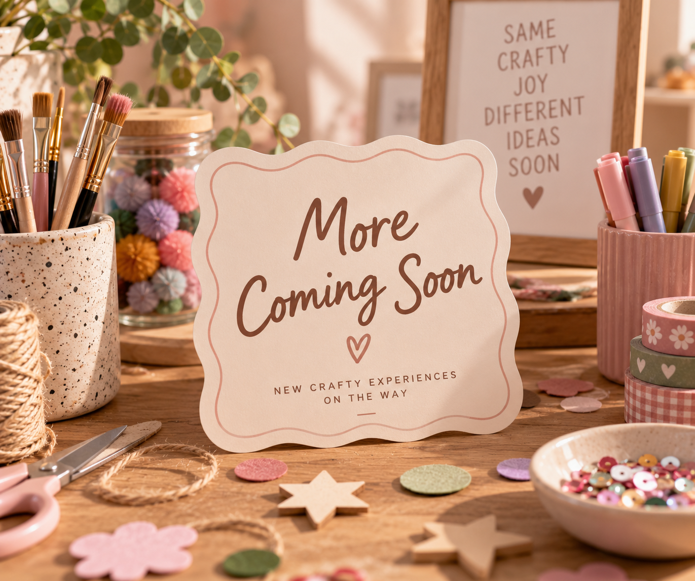 A craft display table with art supplies, colorful pom-poms, and decorative washi tapes, with a sign reading 'More Coming Soon' and a framed message about craft joy.