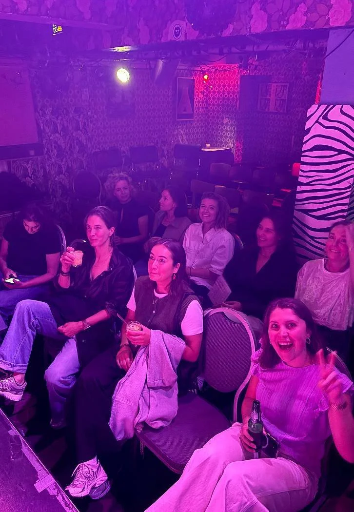 Vrouwelijke Comedy Queens cursisten aanwezigen in een donkere, met roze en paarse lichtverlichting verlichte ruimte, sit- en face-to-face, plezier, drankje in hand, ontspannen sfeer.