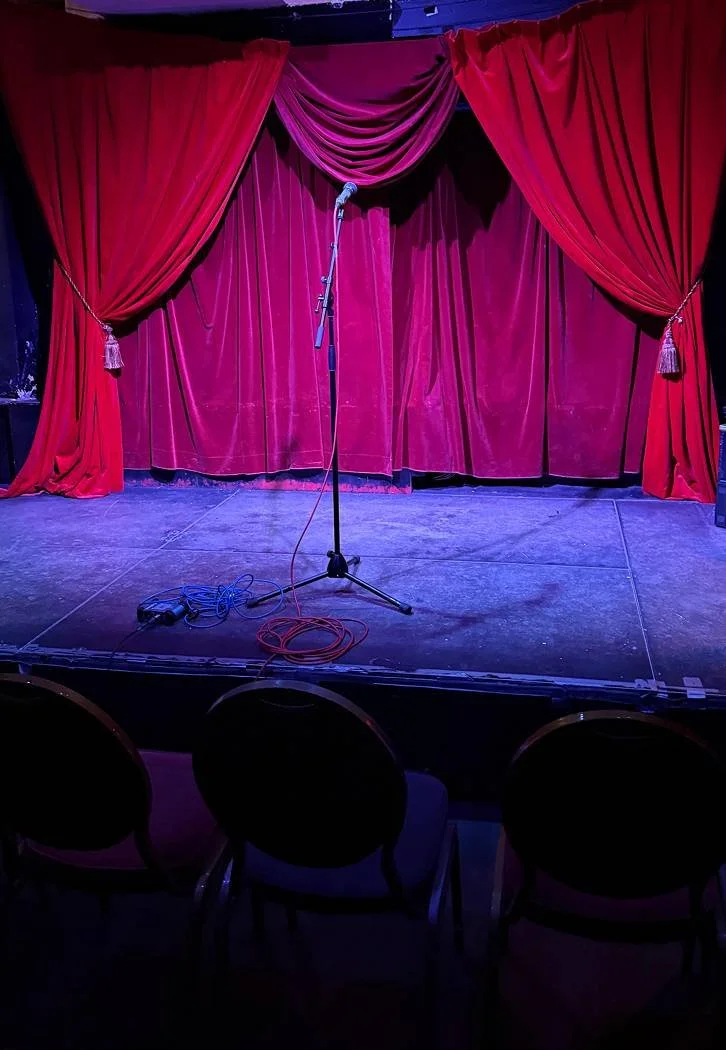 Een podium met een microfoonstandaard, omringd door rode gordijnen met goudkleurige kwasten, klaar voor een optreden of voorstelling.
