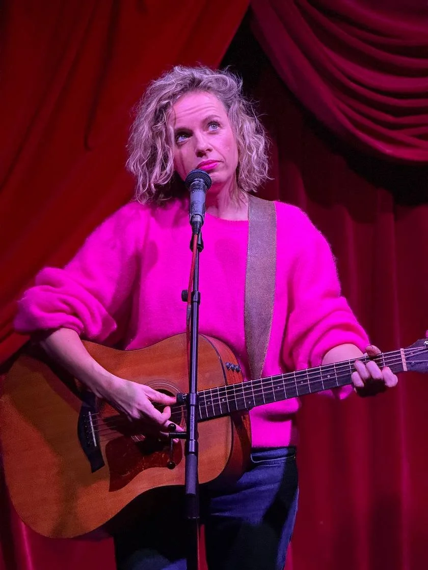 Vrouw zittend op een podium met een gitaar, gekleed in een roze trui, voor een rood fluwelen gordijn, onder een microfoon.