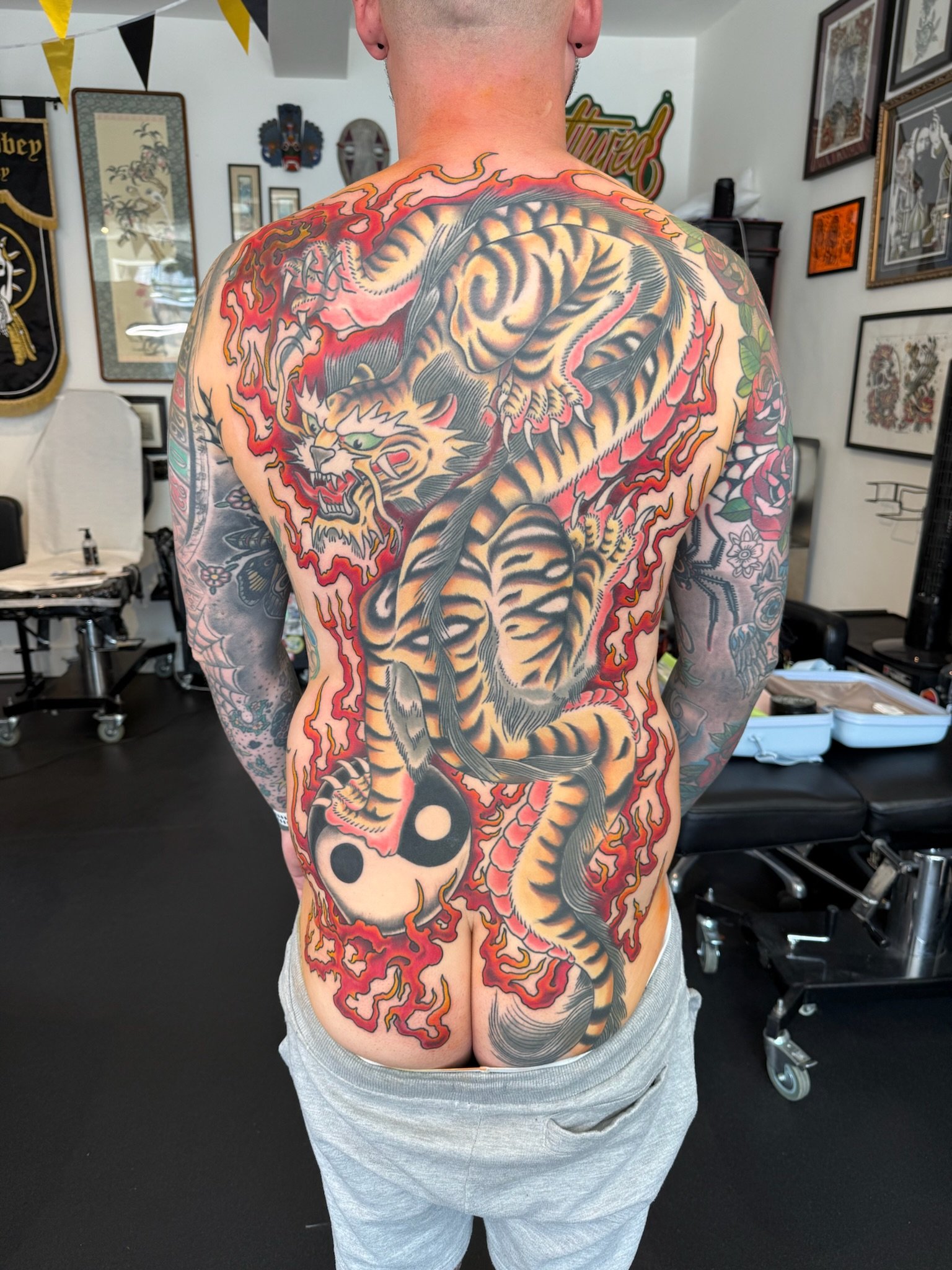 Tattooed person's back with a detailed tiger and dragon tattoo in red, black, and yellow colors, featuring flames and a yin-yang symbol at the bottom.