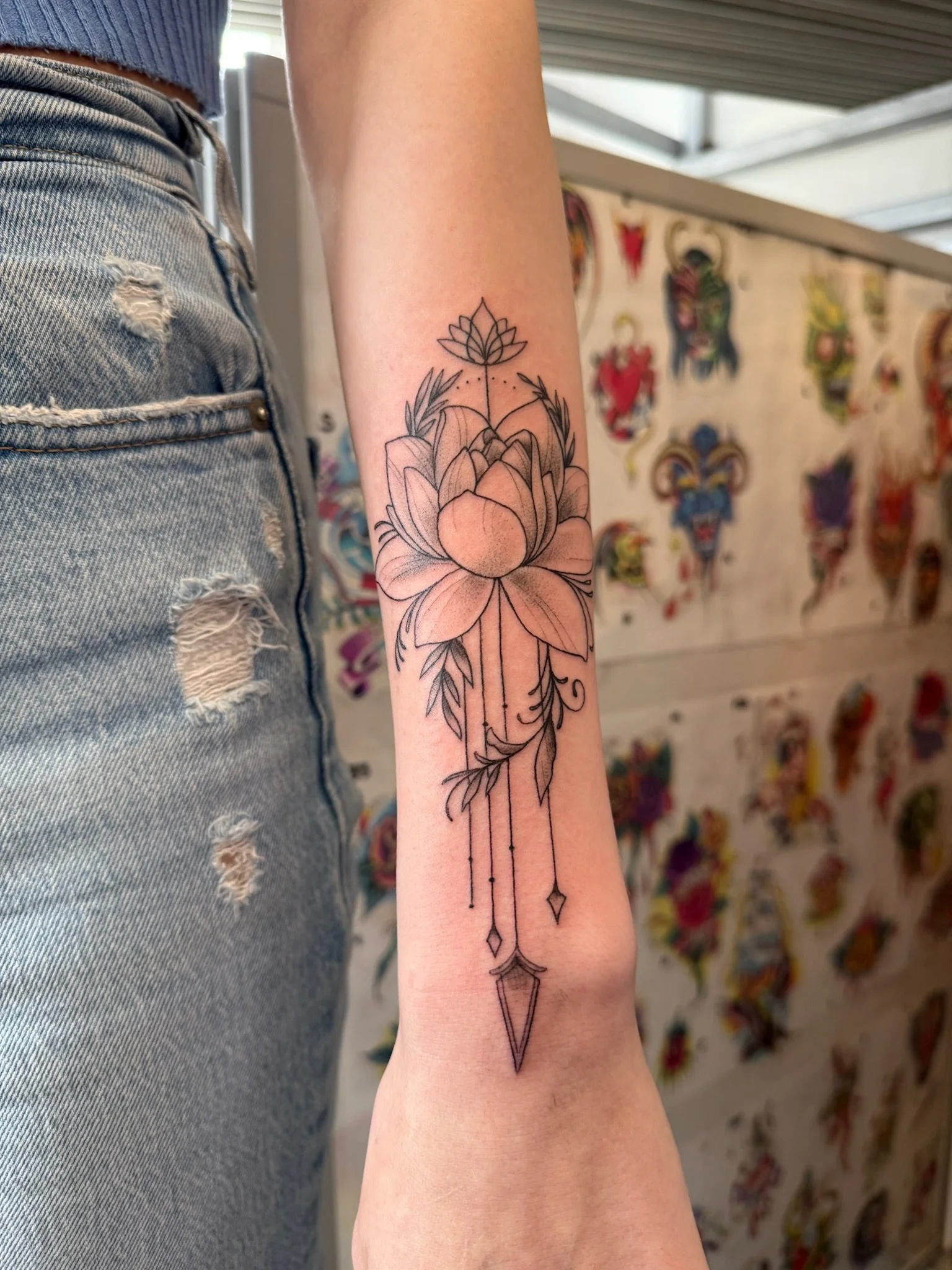A person's arm with a detailed black ink tattoo of a lotus flower at the top, with leaves and a long, ornate arrow pointing downward, all in a minimalistic line art style.