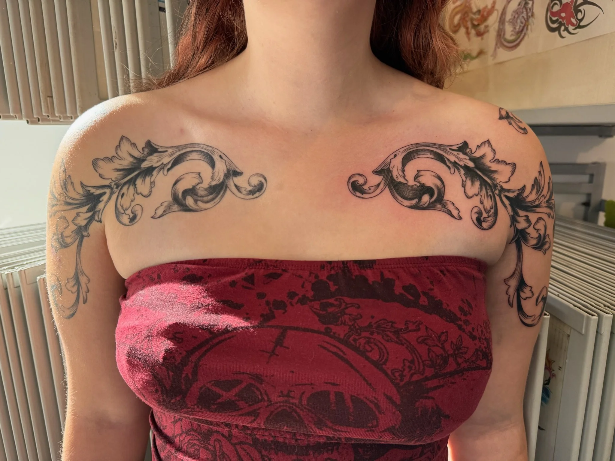 Close-up of a woman’s upper chest and shoulders with a large ornate black tattoo of swirling floral and leaf designs across her collarbones and upper arms. She wears a red strapless top or dress, and her reddish-brown hair is visible on her shoulders. The background includes a heating radiator, some artwork, and other objects.