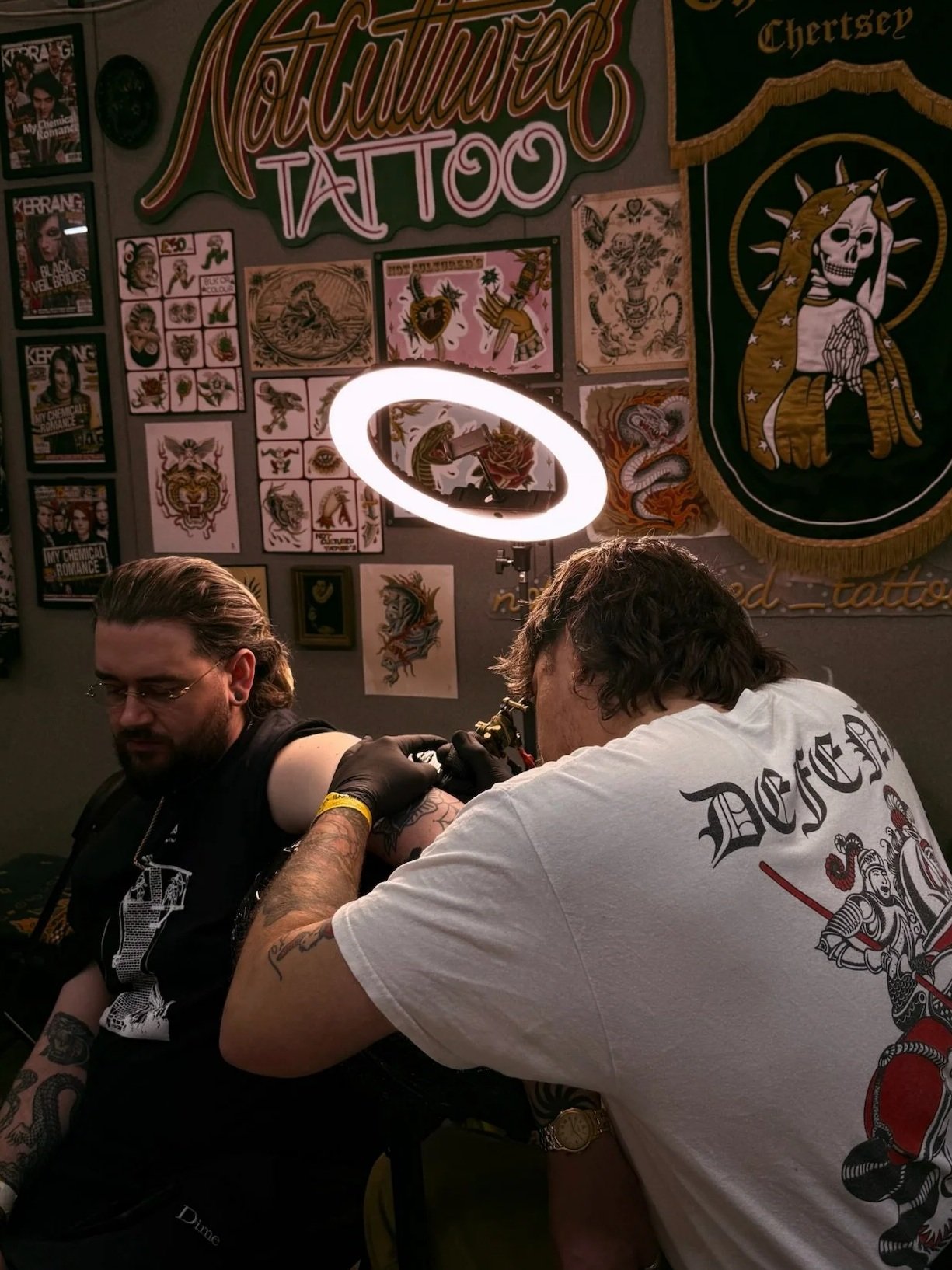 A person receiving a tattoo on their upper arm from a tattoo artist in a tattoo studio. The studio has tattoo-themed artwork and posters on the walls, including a large banner with a skull and snake motif. A ring light shines overhead.