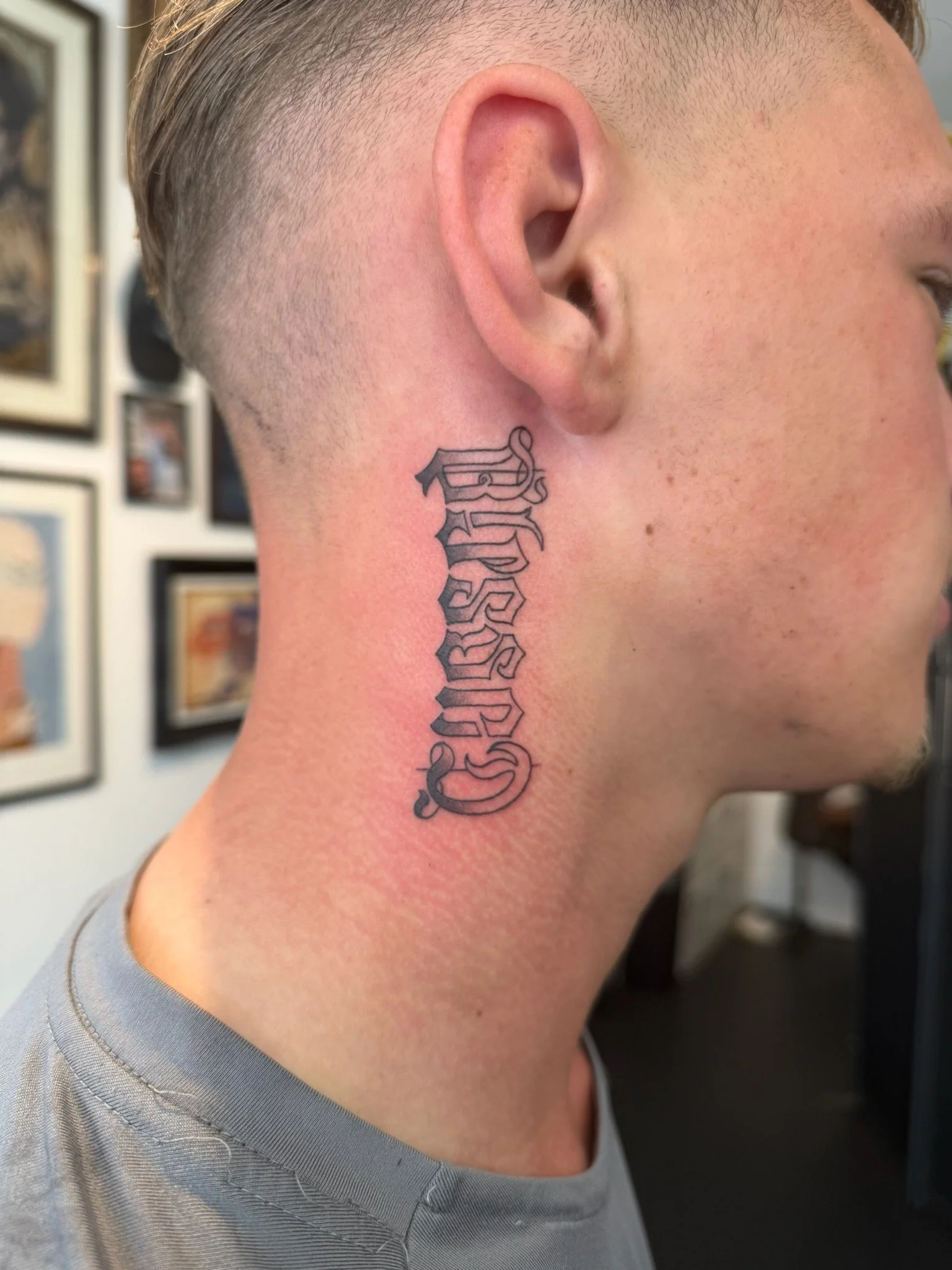 Close-up of a person's left side of the neck with a tattoo of the word 'Sincerely' in Gothic script, with a blurred background showing wall decor.