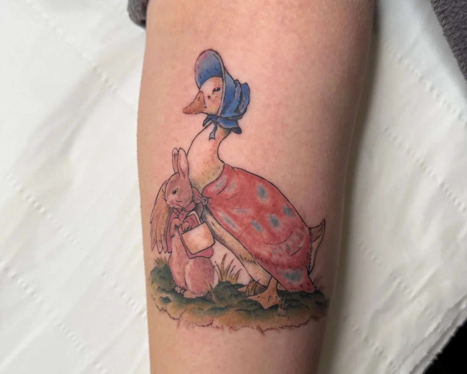 Tattoo of a cartoon duck with a carrot nose and a blue bonnet, holding a rabbit in a pink dress, sitting on a rock.