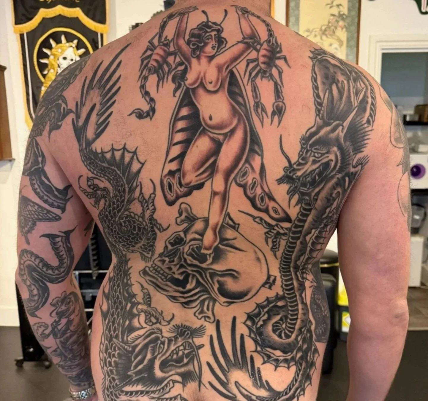 Back tattoo of a woman with horns, wings, and a dress holding a large scorpion, surrounded by a dragon, a wolf, and a snake.