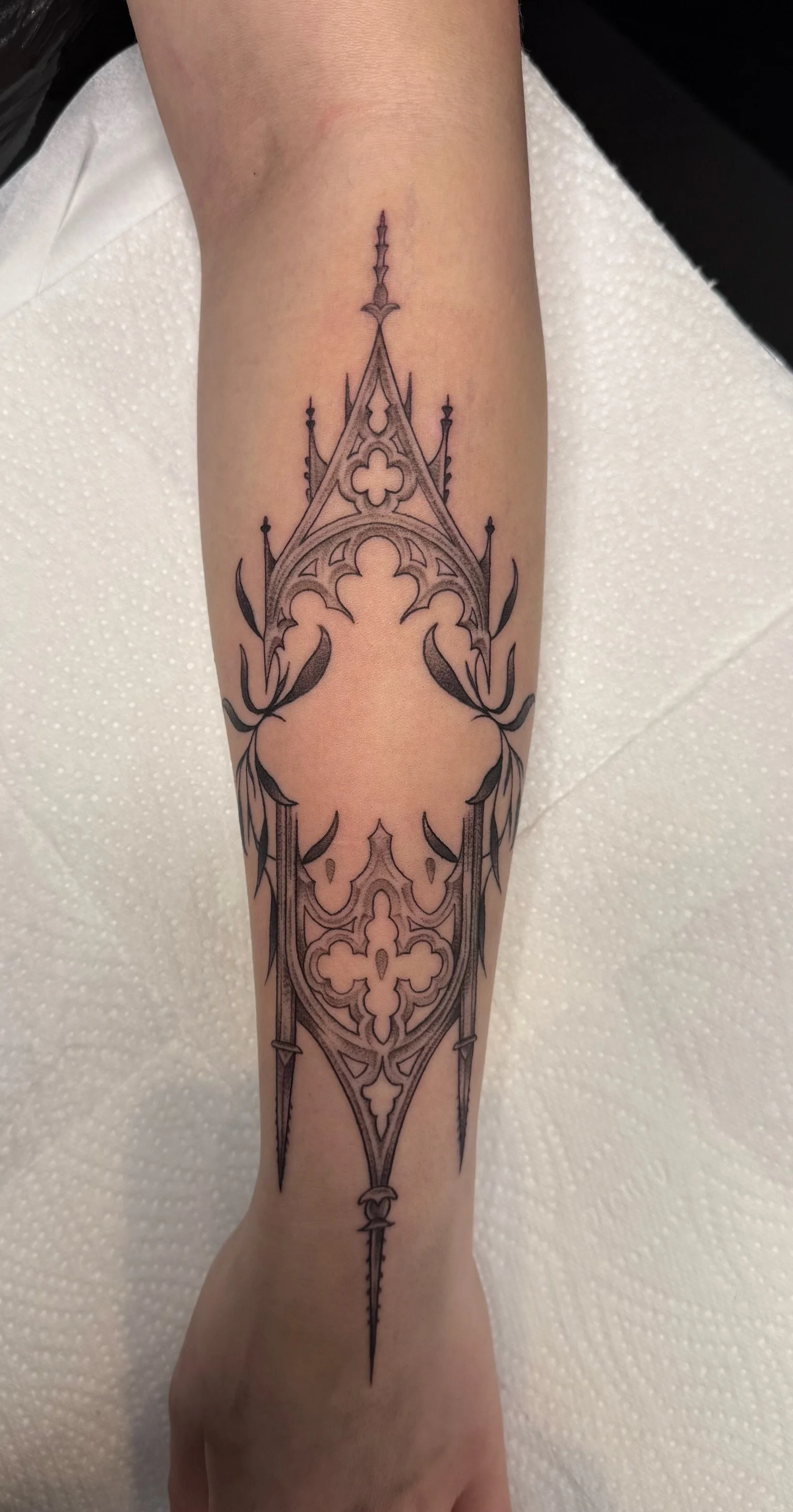 Detailed black line tattoo on forearm resembling gothic cathedral architecture with intricate ornamental designs and pointed spires.