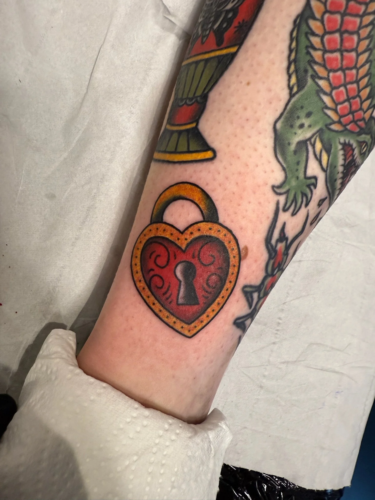 Tattoo of a heart-shaped padlock on forearm, surrounded by colorful floral and dinosaur-themed tattoos.