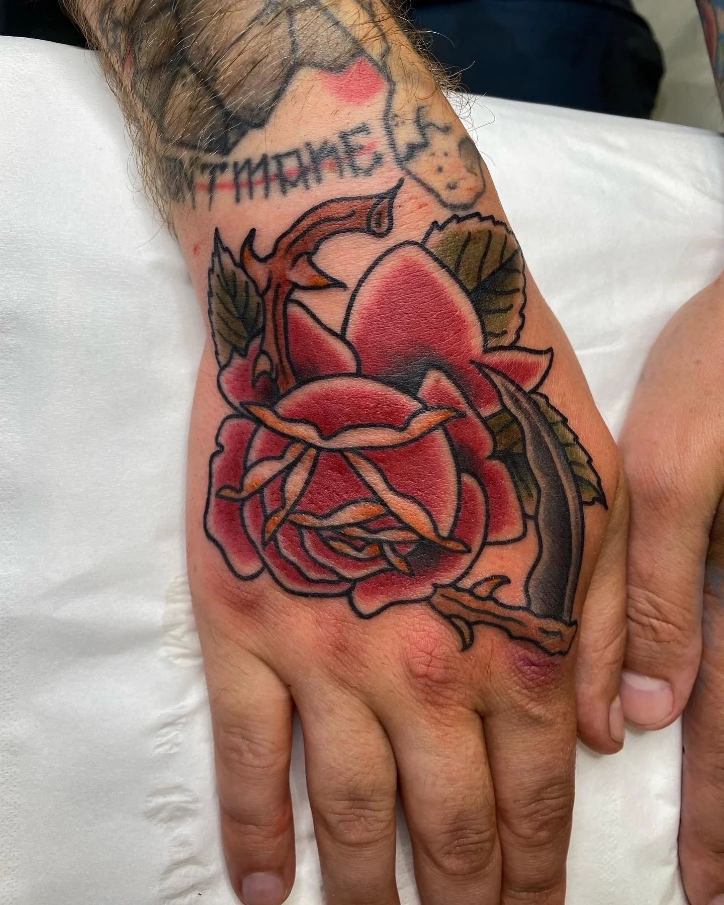 A tattoo on a person's hand features a red and black rose with green leaves. There is also a partial tattoo of text above the rose that reads 'TREATMENT'.