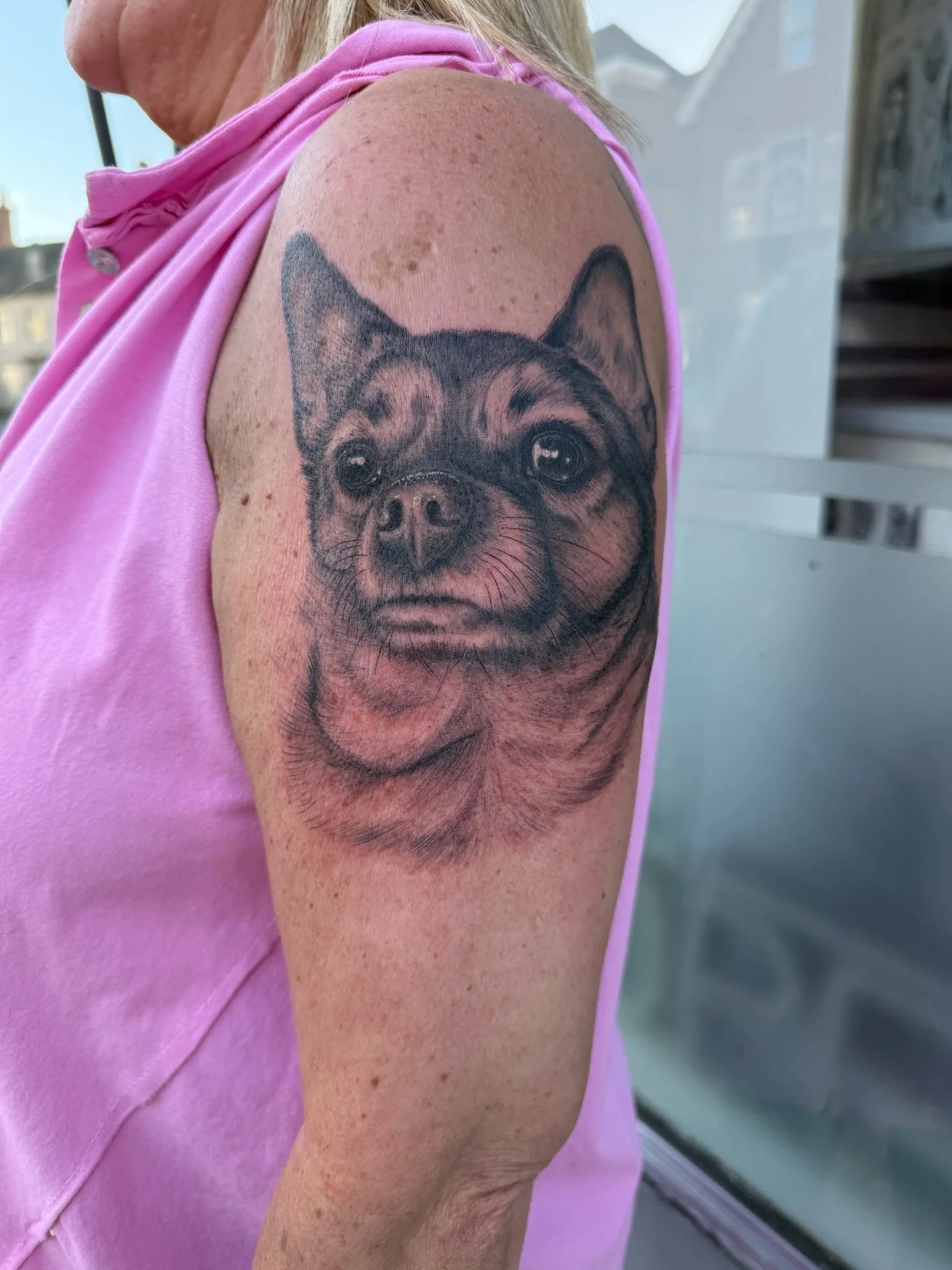 Close-up of a person's upper arm showing a detailed black and gray tattoo of a dog's face, possibly a Chihuahua, with a pink sleeveless top and pink scarf. Background includes part of an outdoor or industrial setting.